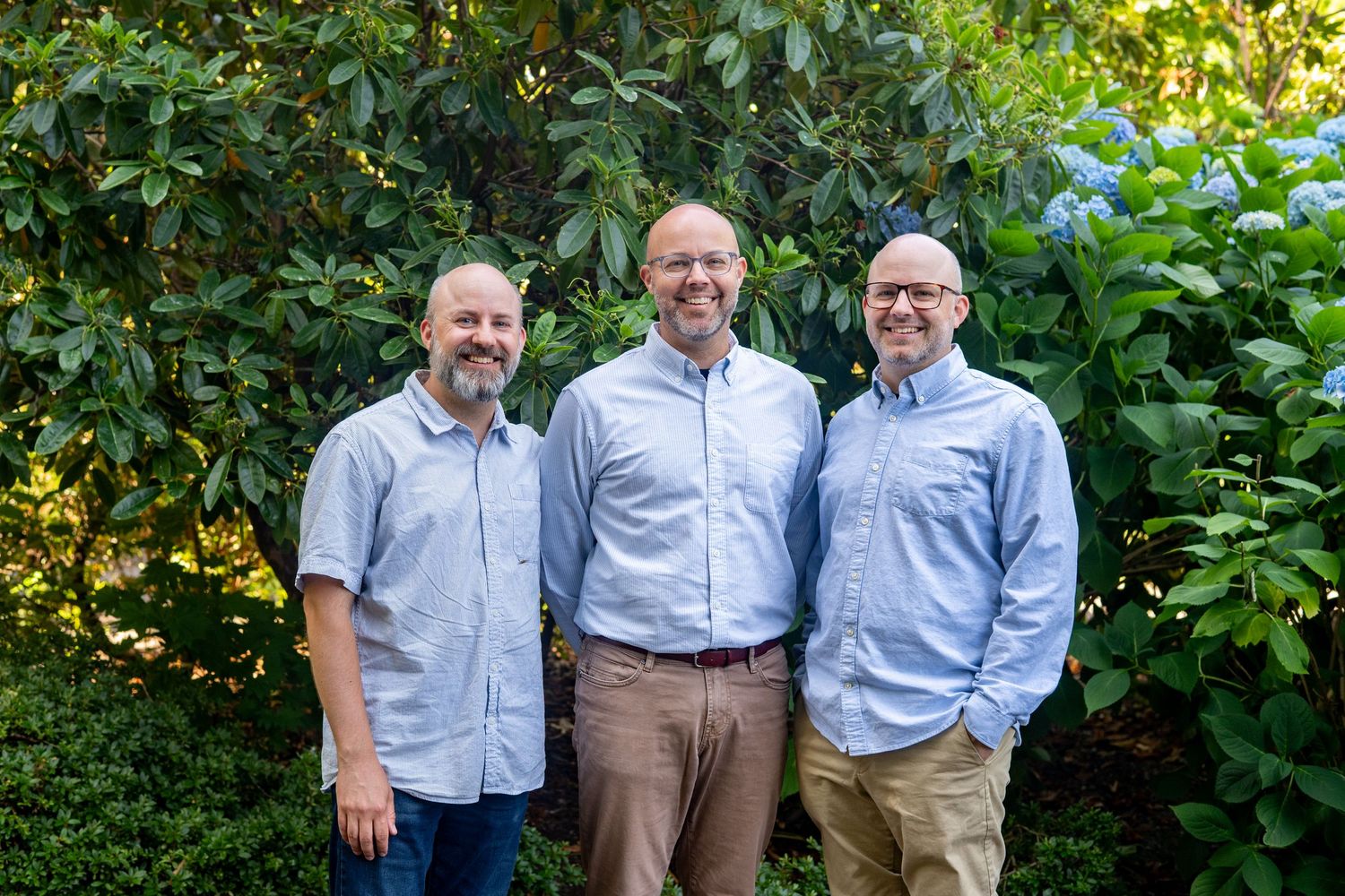 Three people in light blue dress shirts and khaki pants pose together in a lush green garden setting.