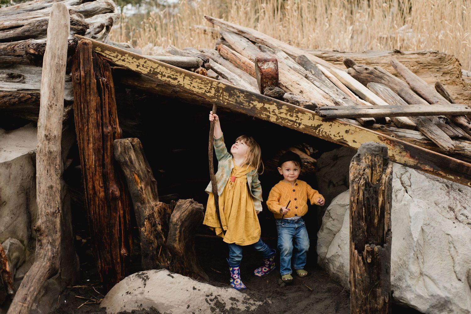 Two children exploring a rustic wooden fort made of sticks and logs in nature.