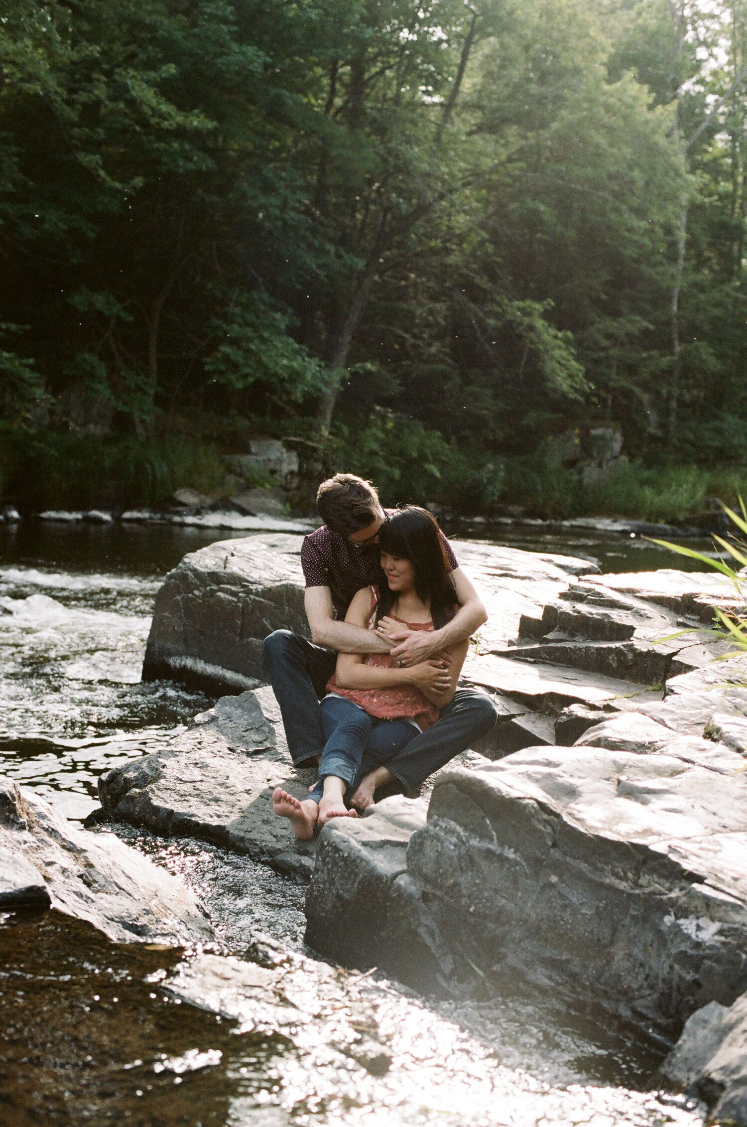 A couple sits peacefully together on rocks by a flowing stream in a shaded forest setting.