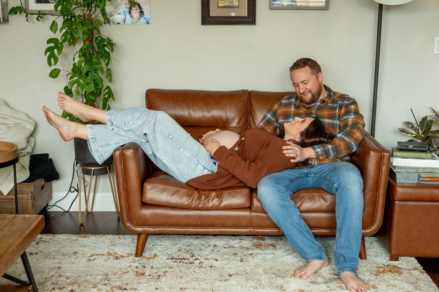 Pregnant couple relaxing comfortably on a brown leather couch in a bright living room with plants and decor.