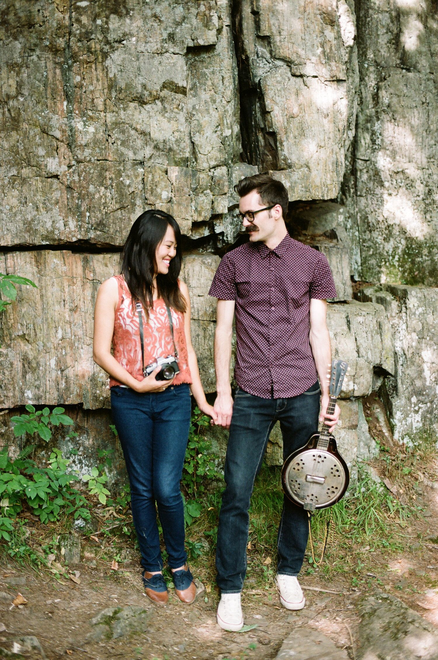 A couple stands together against a stone wall backdrop as they check their mobile devices.