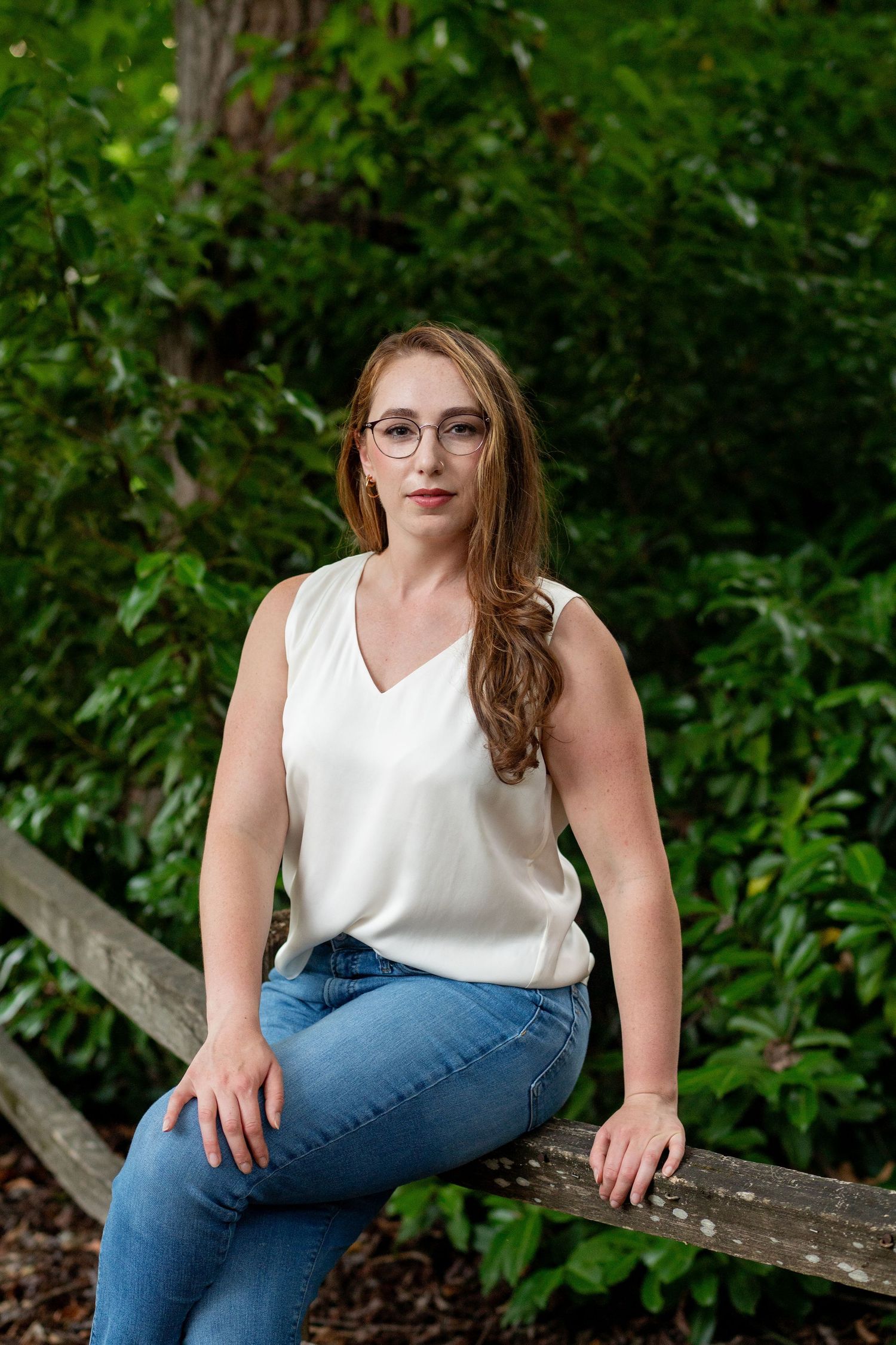 A person in glasses, white sleeveless top and blue jeans sits casually on a rustic wooden bench against lush greenery.