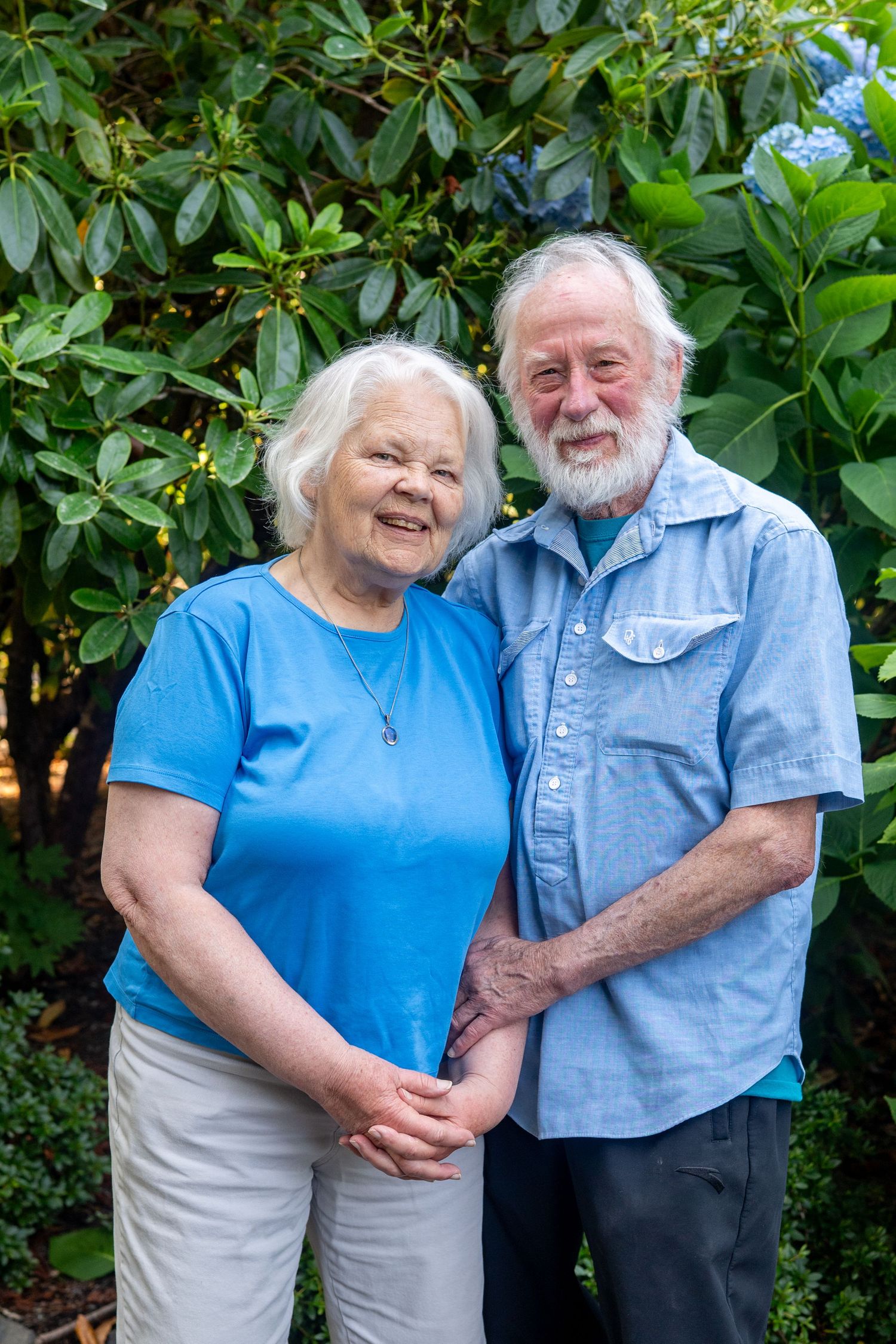 Happy elderly couple in matching blue shirts embrace and smile outdoors in front of green foliage.