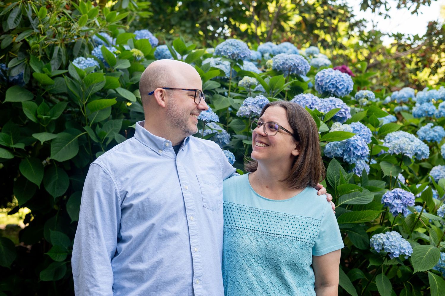 Couple stands together in front of blooming blue hydrangea bushes in a garden setting.