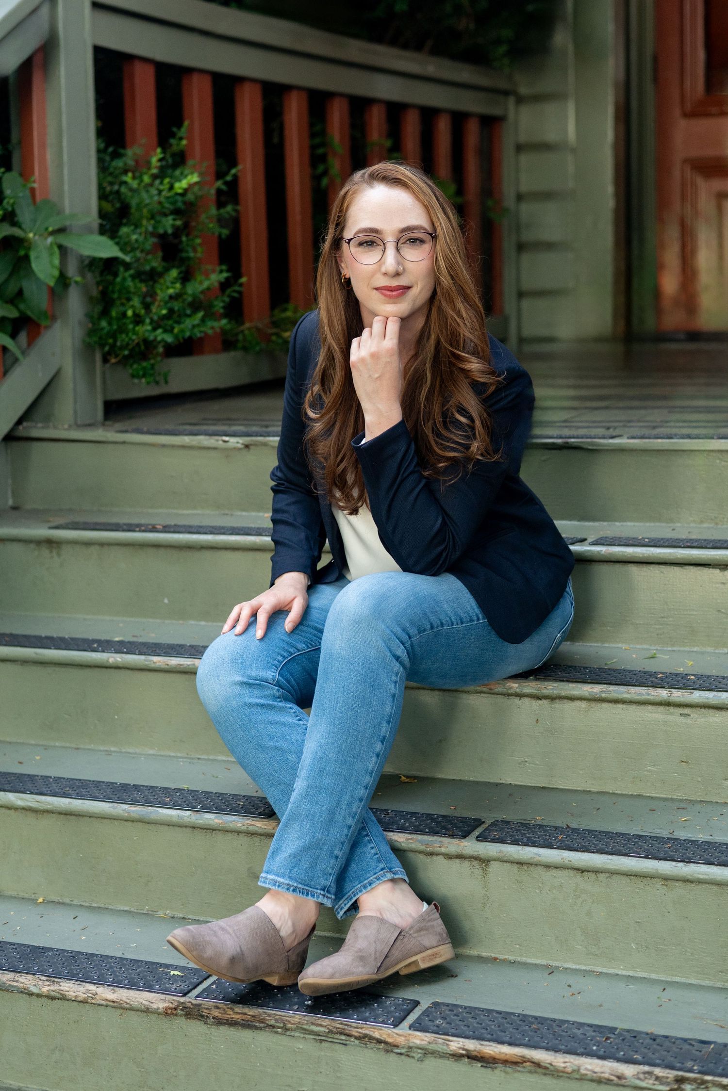 A person wearing blue jeans and brown shoes sits casually on outdoor wooden steps near a building entrance.