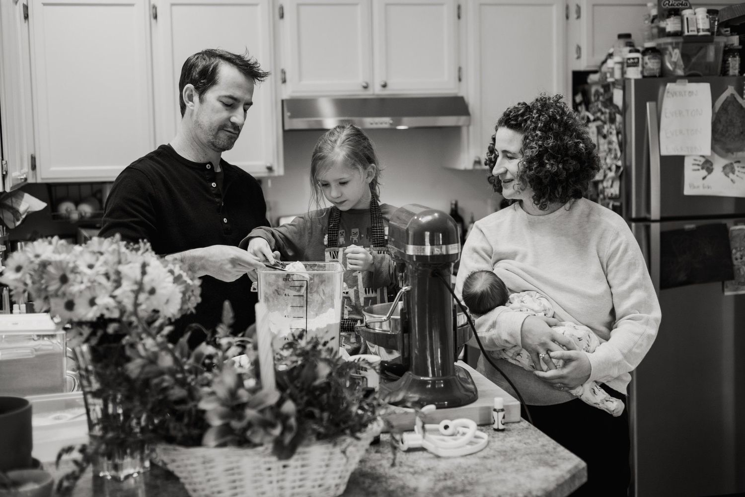 Three people gathered in a kitchen around a floral centerpiece and drinks in a black and white photograph.
