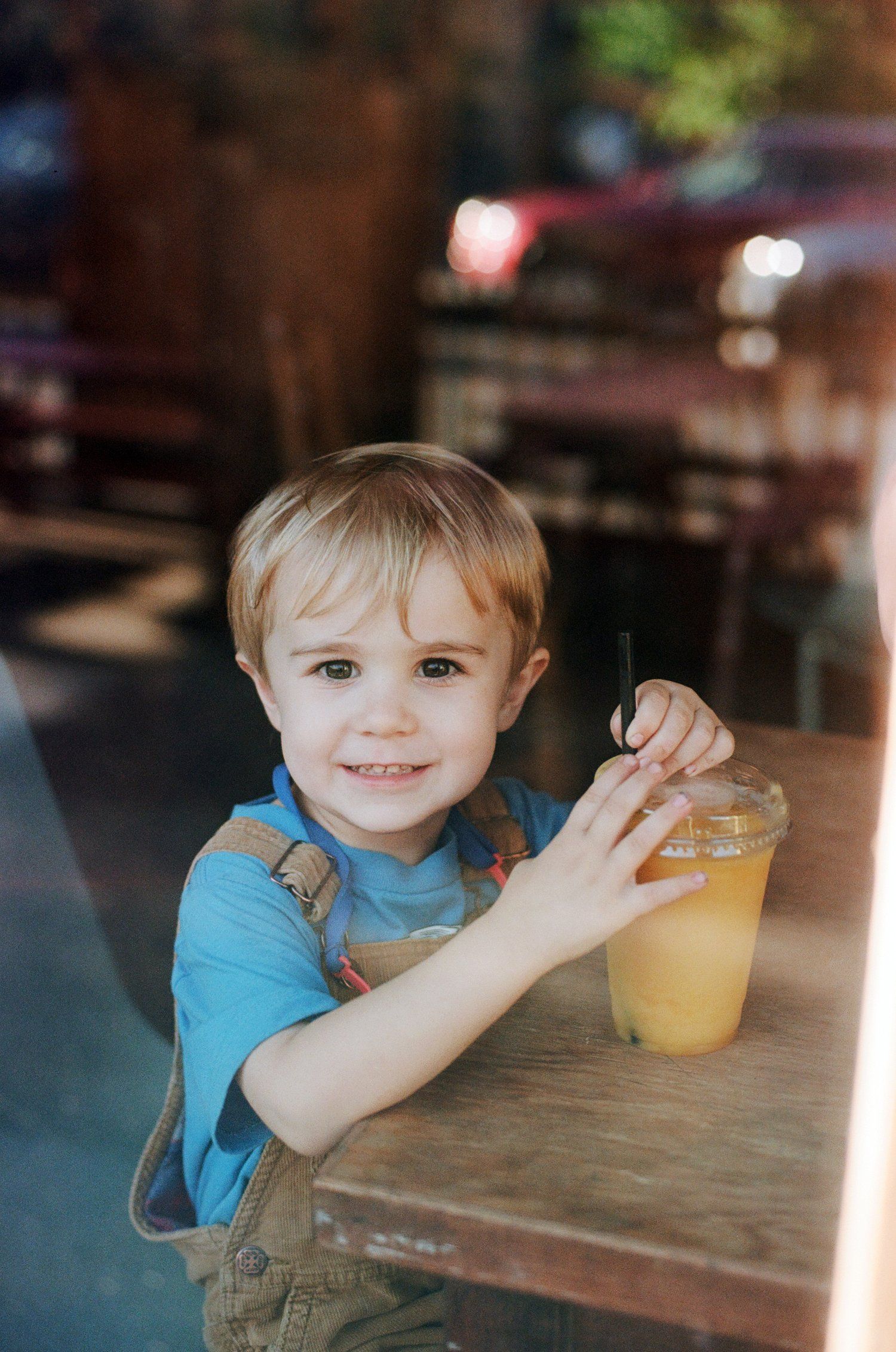 A child in blue shirt sits at wooden table enjoying a beverage from a clear plastic cup.