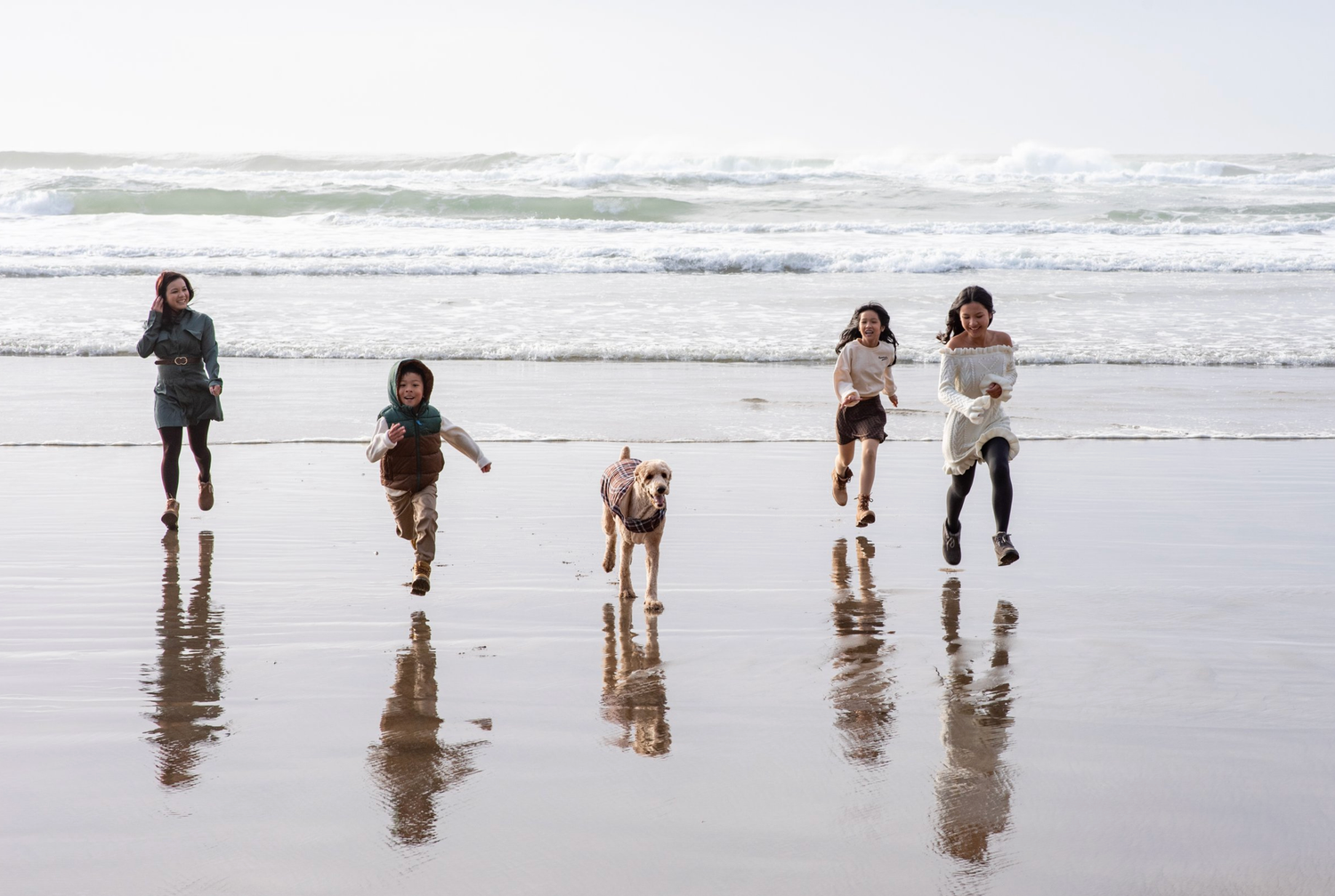 Group running and playing with dog along reflective beach shoreline at sunset.