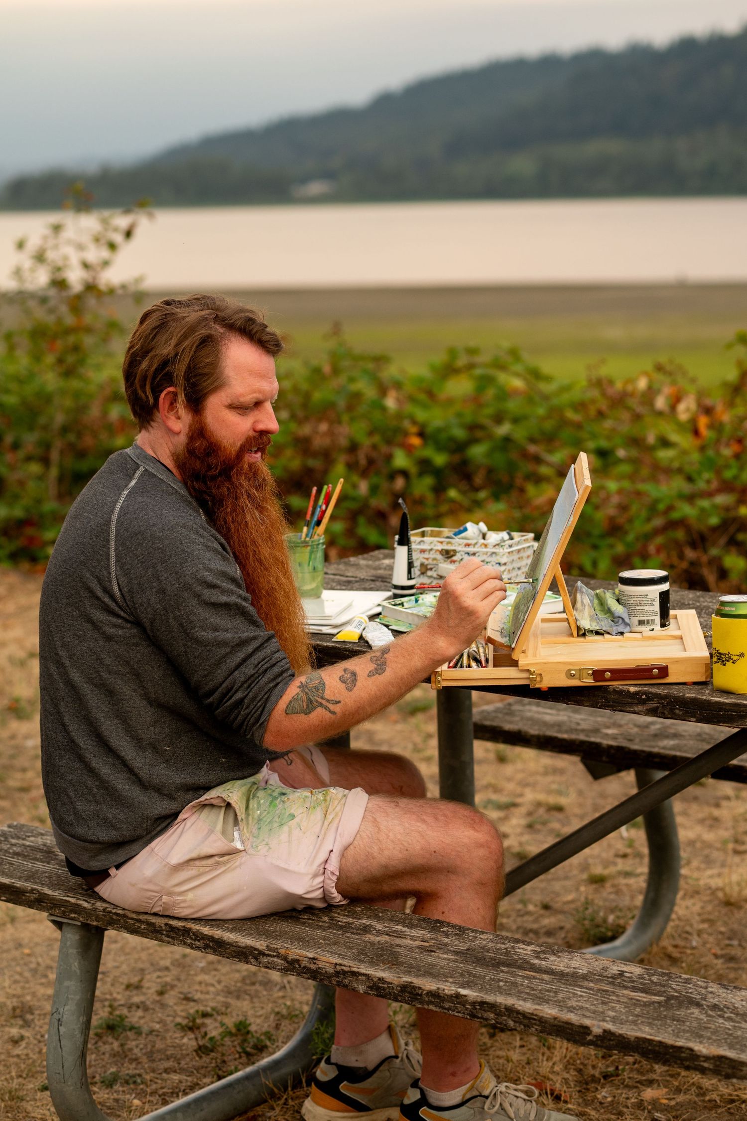 A bearded man in a gray shirt sits at a wooden picnic table near a beach, enjoying food and drink at sunset.