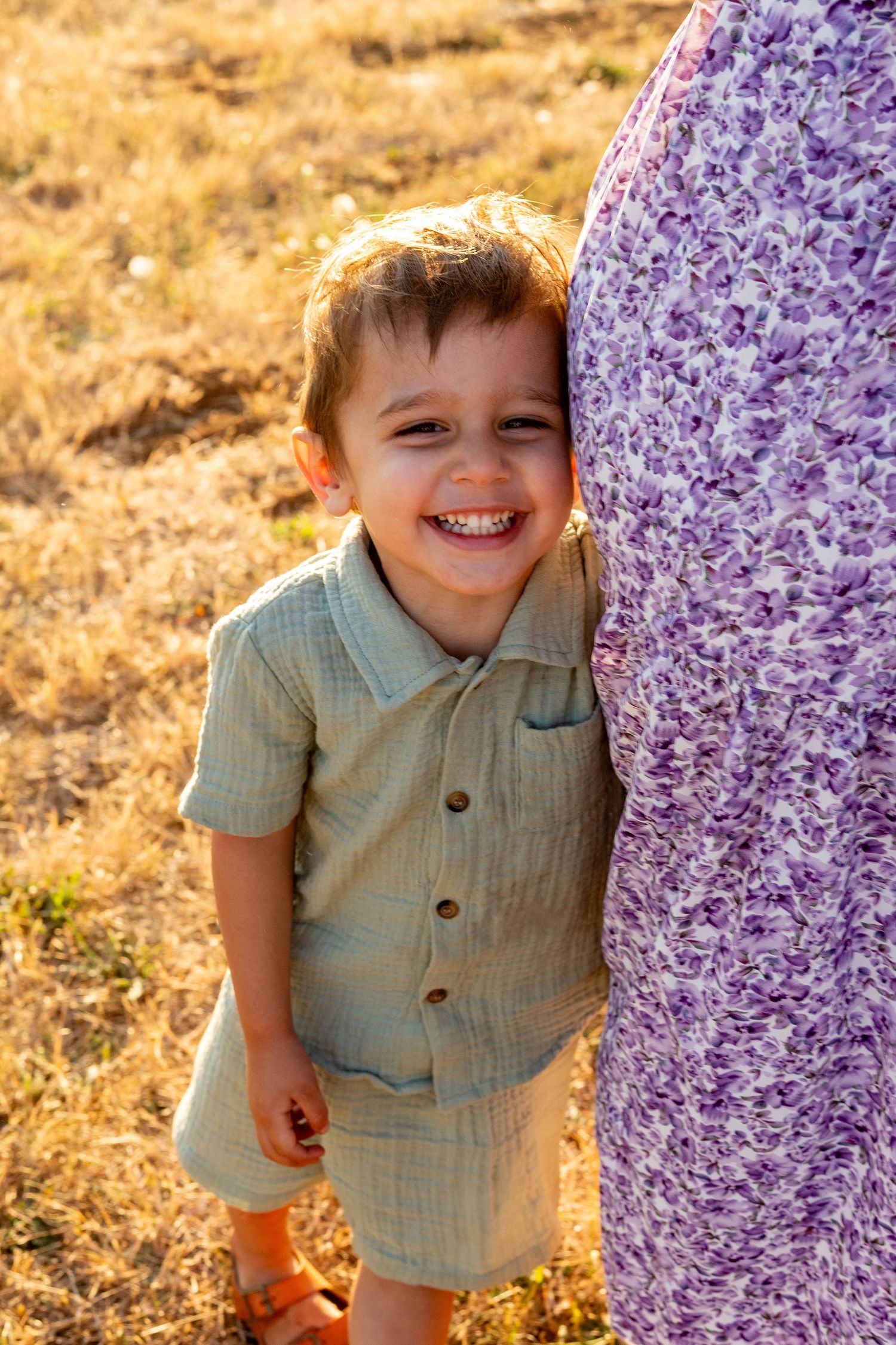 Small child in mint green outfit peeks out playfully from behind purple floral fabric in golden sunlit setting.