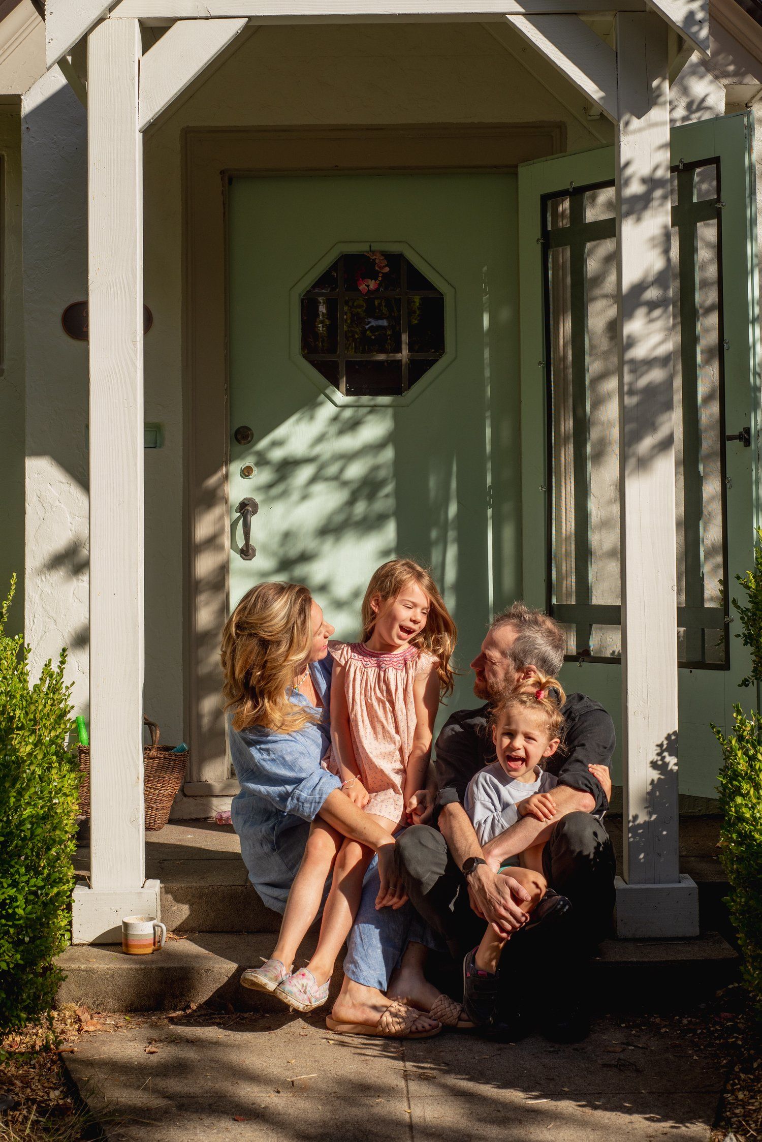 A family sits together on a front porch step with shadows from a tree casting dappled light on a mint green door.