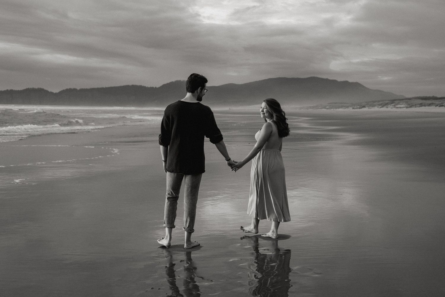 A couple holding hands while standing in shallow ocean waves on a cloudy beach.