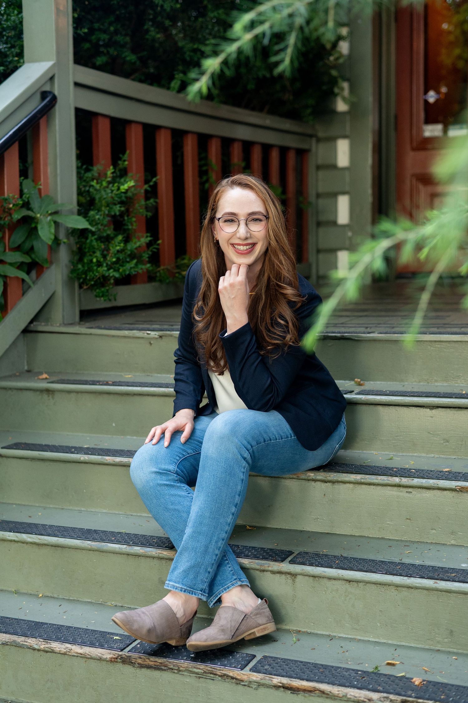 A person wearing glasses and jeans sits casually on outdoor concrete steps near greenery and wooden railings.