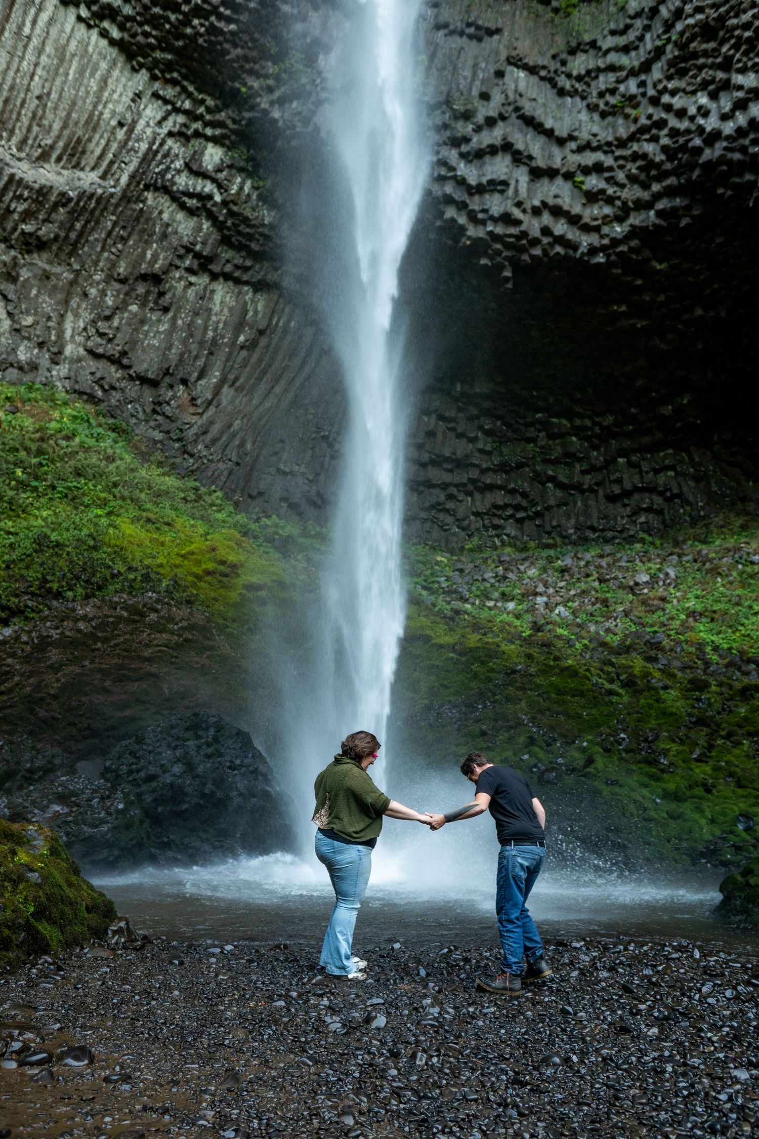 Two people stand together near the base of a tall, misty waterfall surrounded by mossy rock faces and gravel.