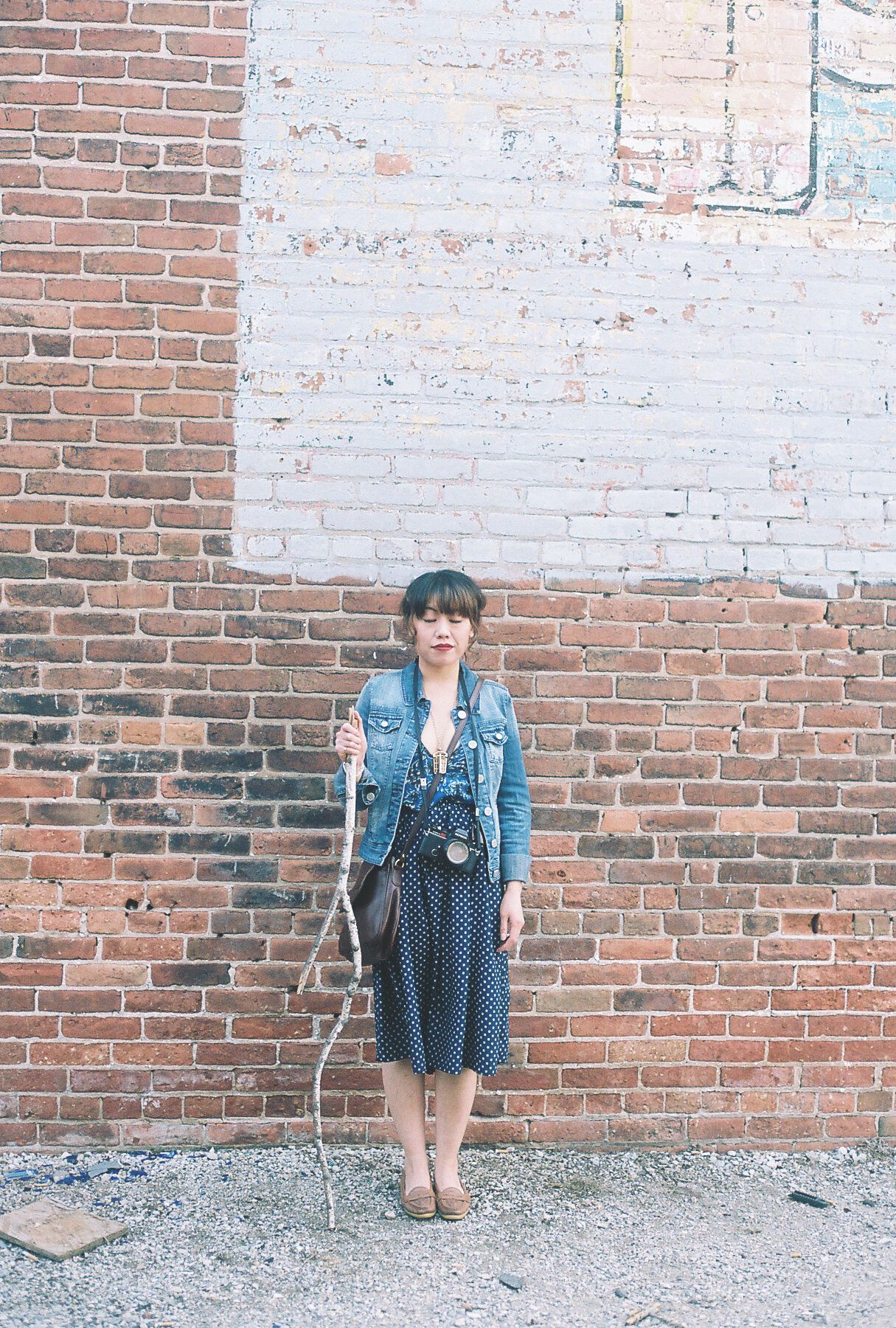 Someone wearing a denim jacket and polka dot dress poses against a weathered brick wall with vintage camera.