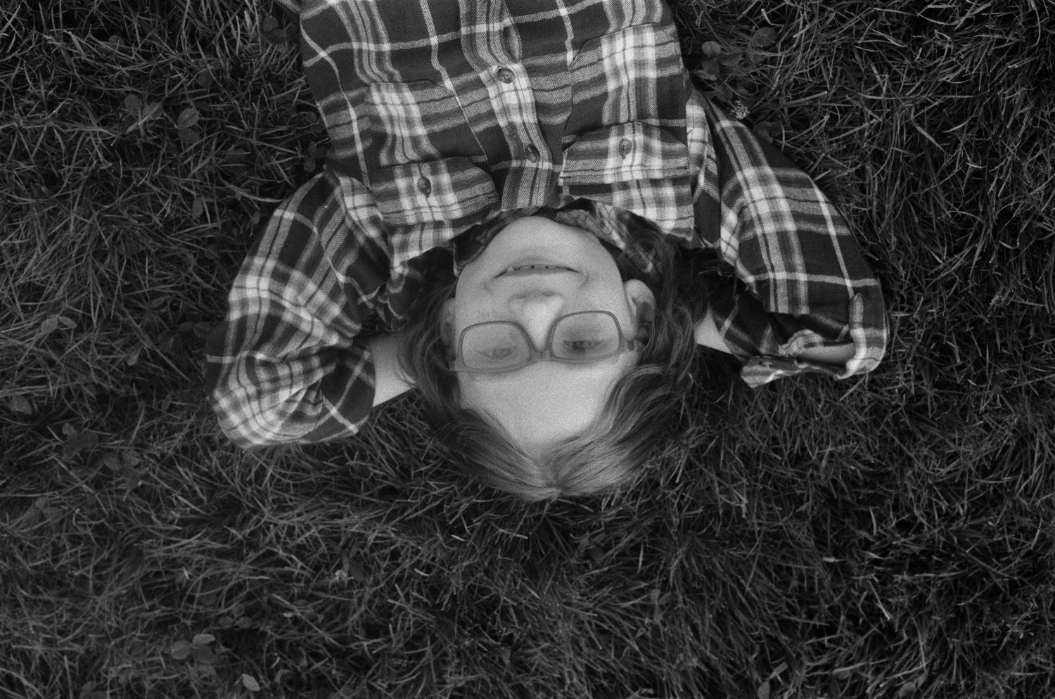 Black and white overhead view of someone wearing glasses lying in grass wearing a plaid shirt.