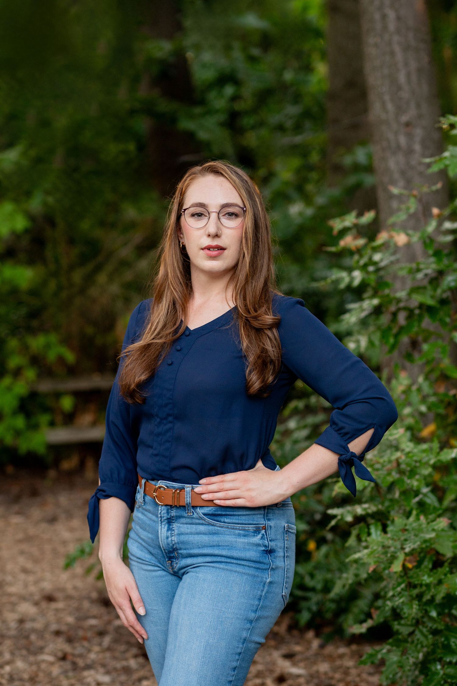 A person in a navy blue top and light denim jeans poses outdoors against a green leafy background.