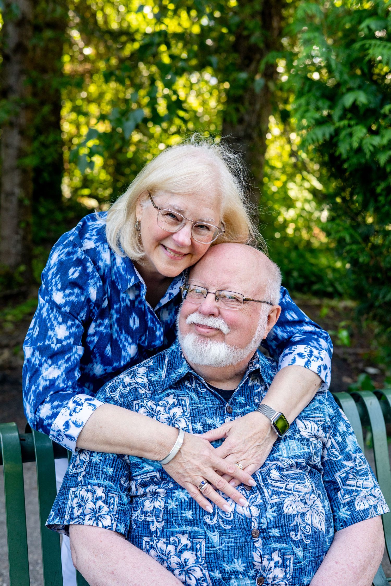 Couple wearing matching blue floral shirts embrace lovingly on a park bench in dappled sunlight.