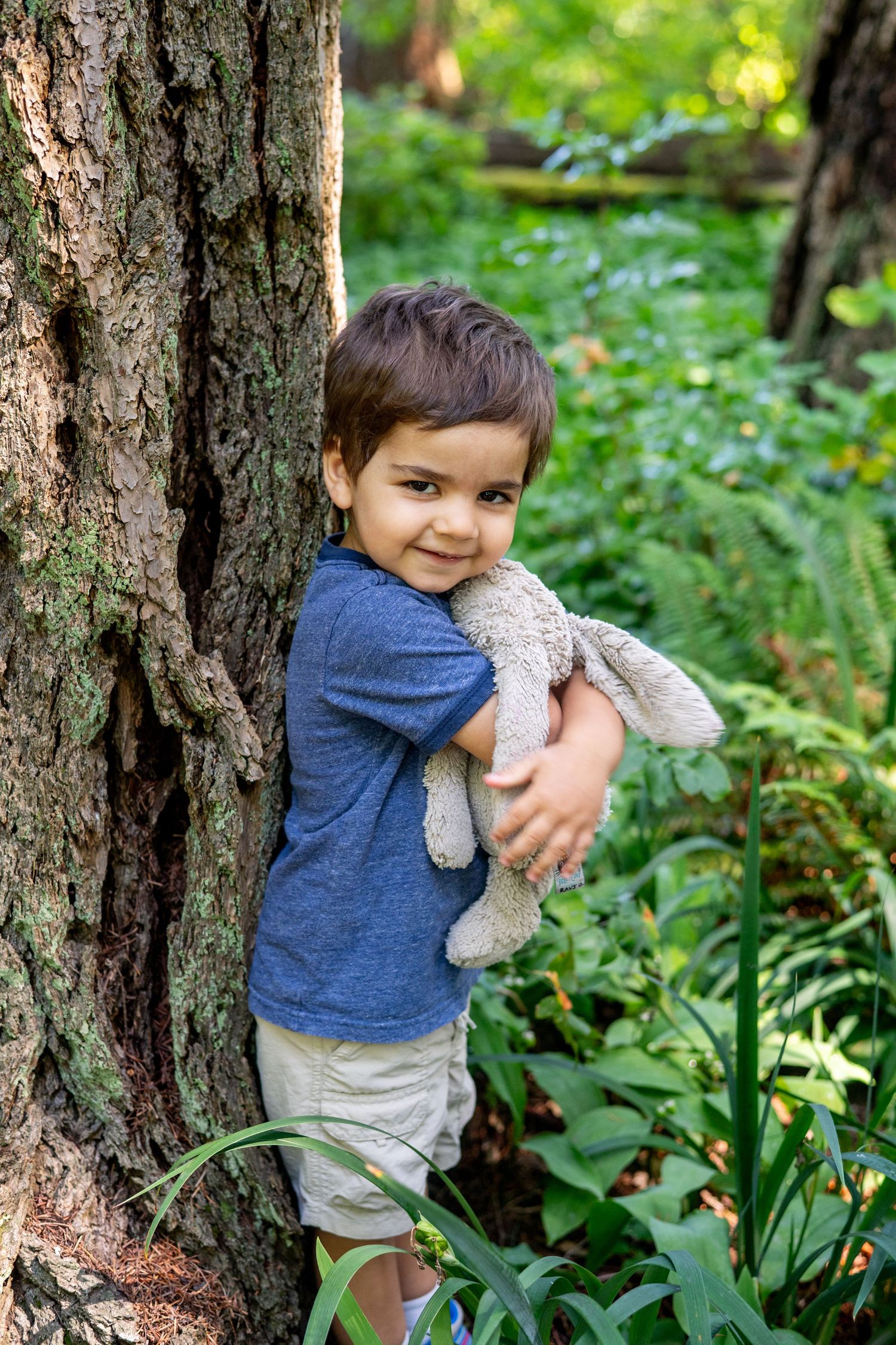 A child in a blue shirt hugs a stuffed animal while standing next to a tree trunk in a green forest setting.