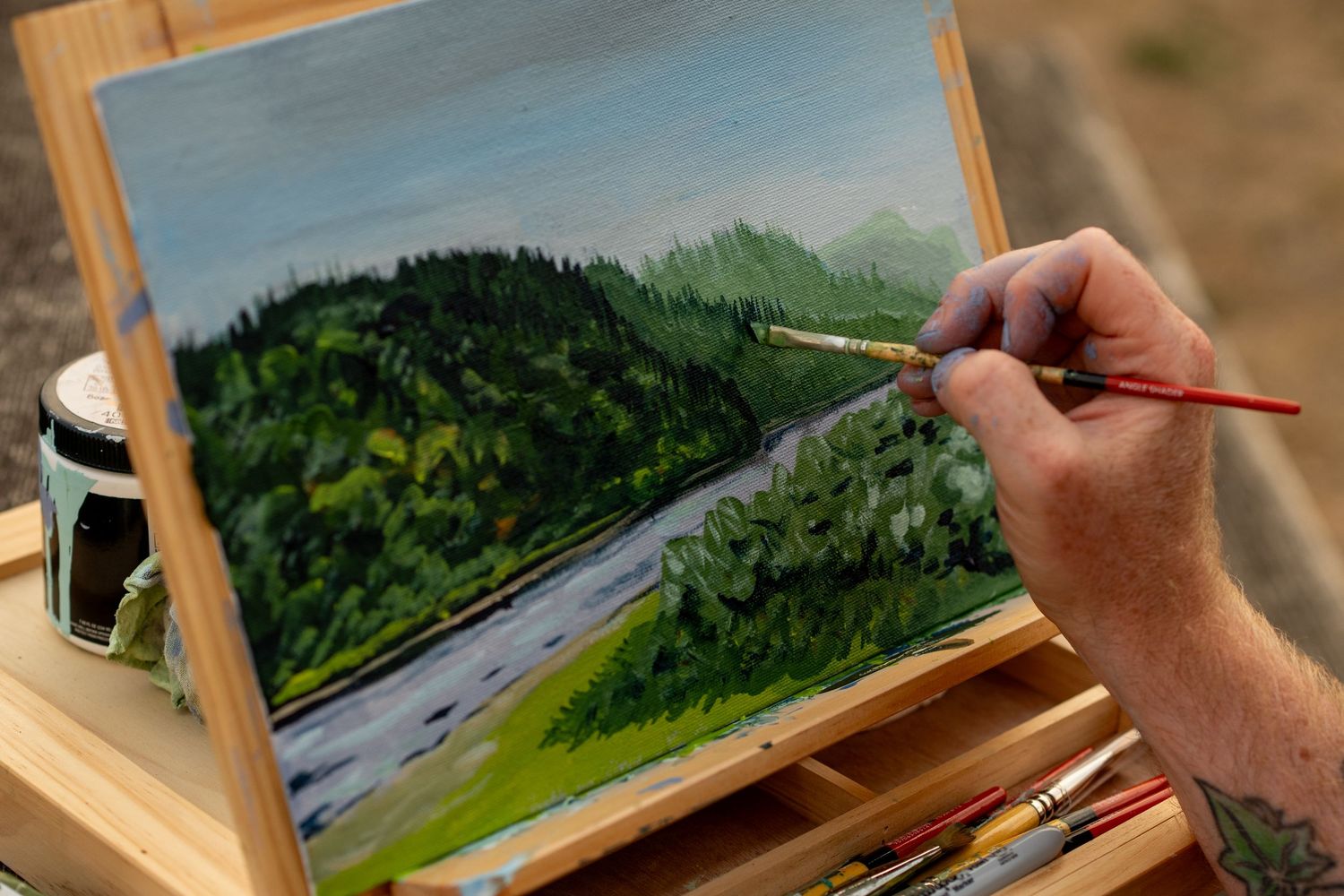 Artist hand painting a detailed landscape mural on a wall depicting a winding mountain highway near forests.