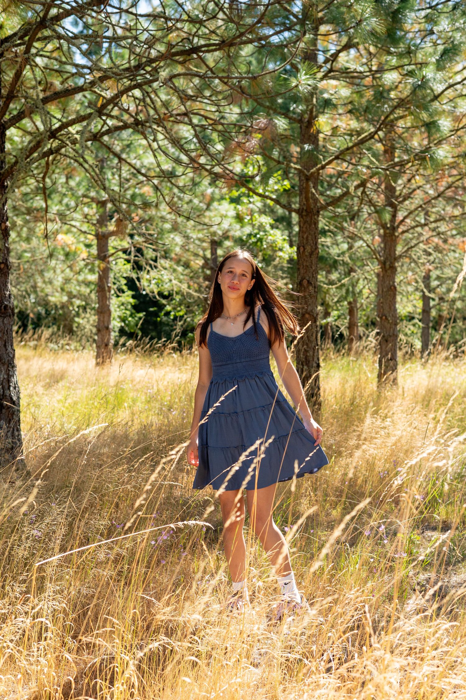 A person in a navy blue dress stands among tall golden grass and pine trees on a sunny summer day.