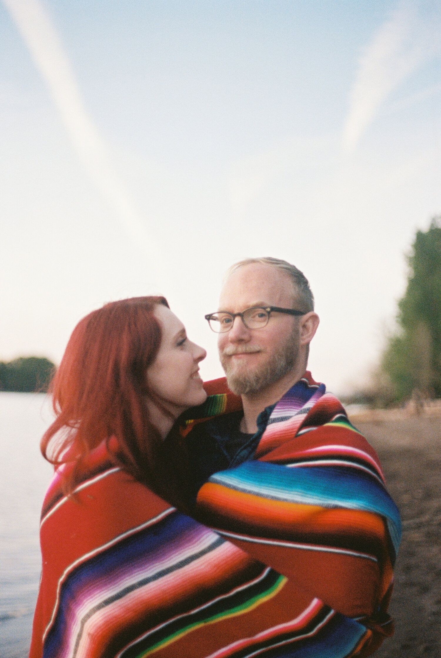 Couple wrapped in colorful striped blanket share intimate moment at sunset by the water.