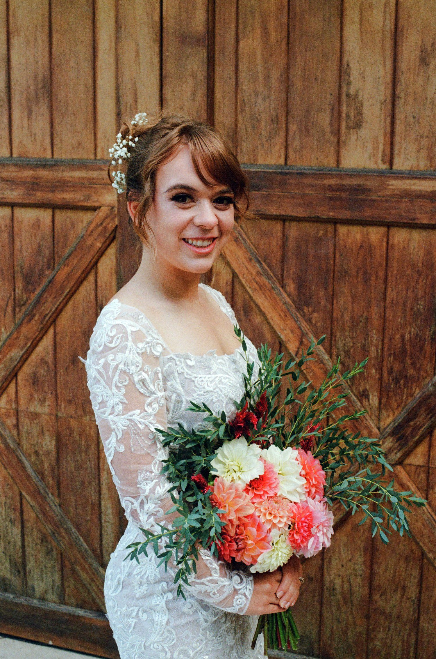 A lace-clad figure holds a vibrant coral and white bouquet against a rustic wooden barn door background.
