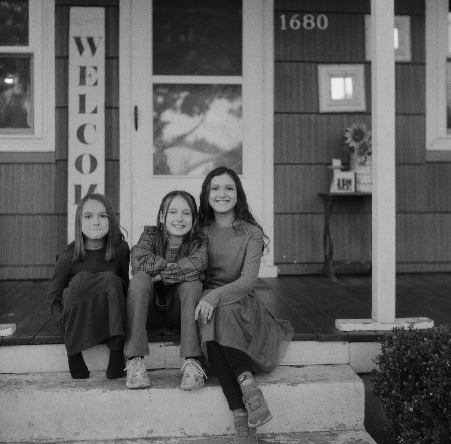 Three people sit on front porch steps of a house with a welcome sign in black and white photograph.