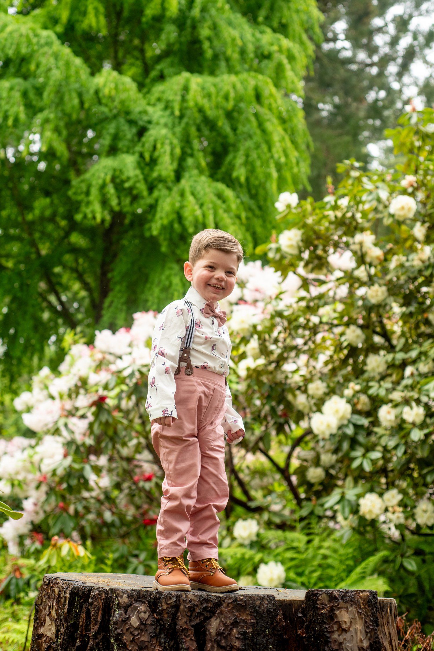 A cheerful child wearing pink overalls stands proudly on a tree stump in a blooming garden.