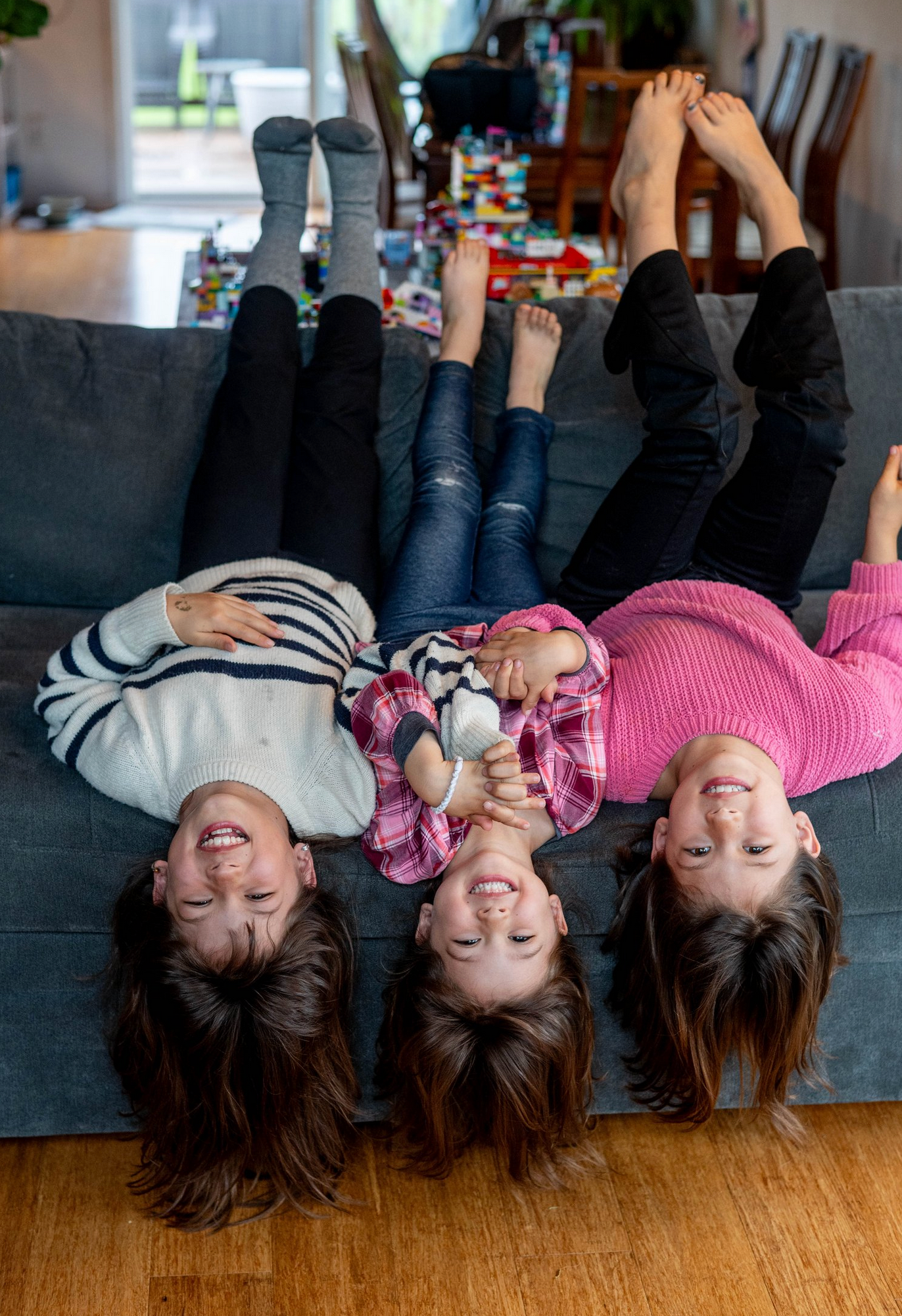 Three people lying upside down on a gray couch with their hair hanging down while laughing and smiling.