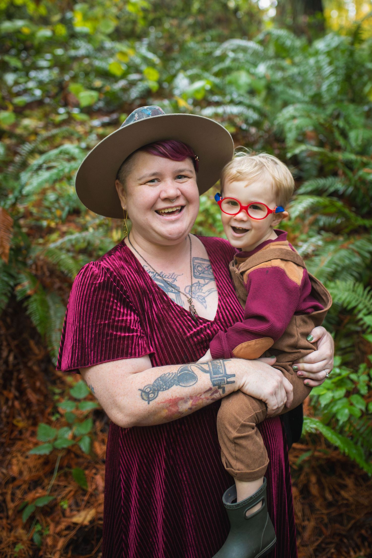 A person wearing a burgundy velvet dress and wide-brimmed hat holds a small child in orange glasses against a forest backdrop.