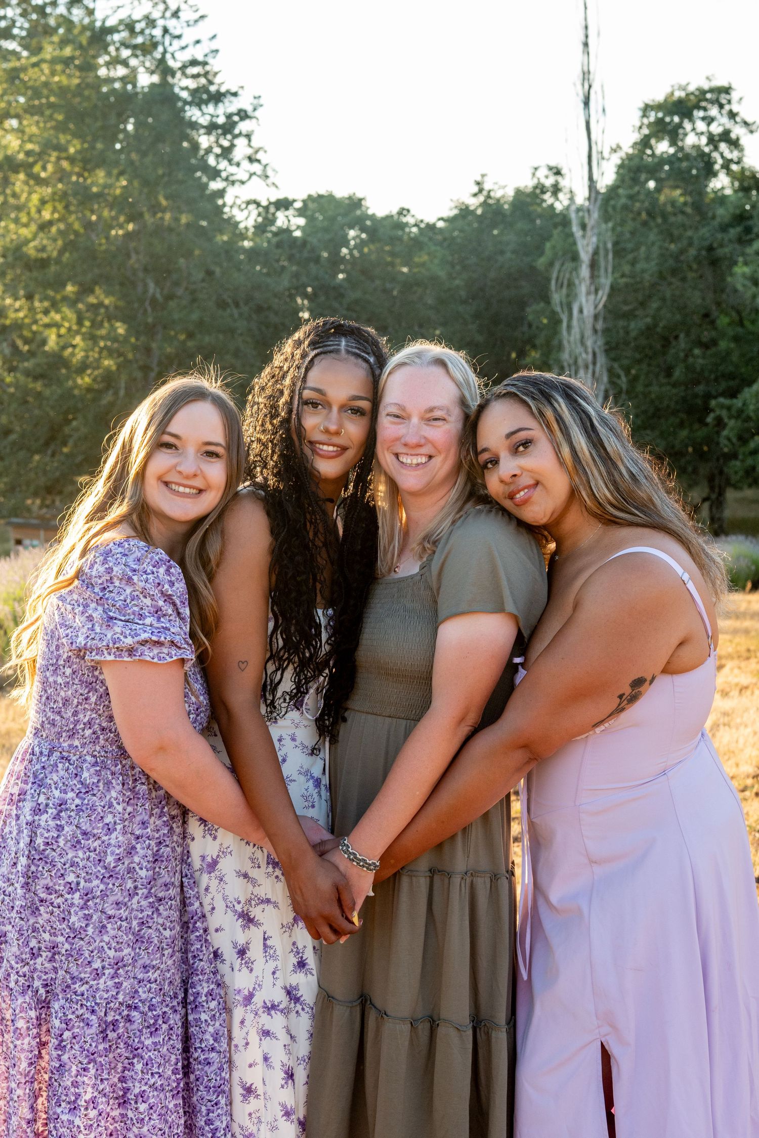A group of four friends in elegant dresses pose together outdoors during golden hour on a summer evening.