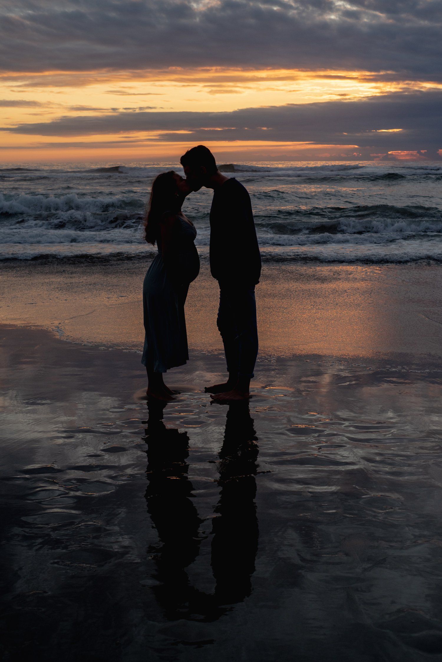 Silhouetted couple sharing an intimate moment on beach at sunset with dramatic orange sky and ocean waves.