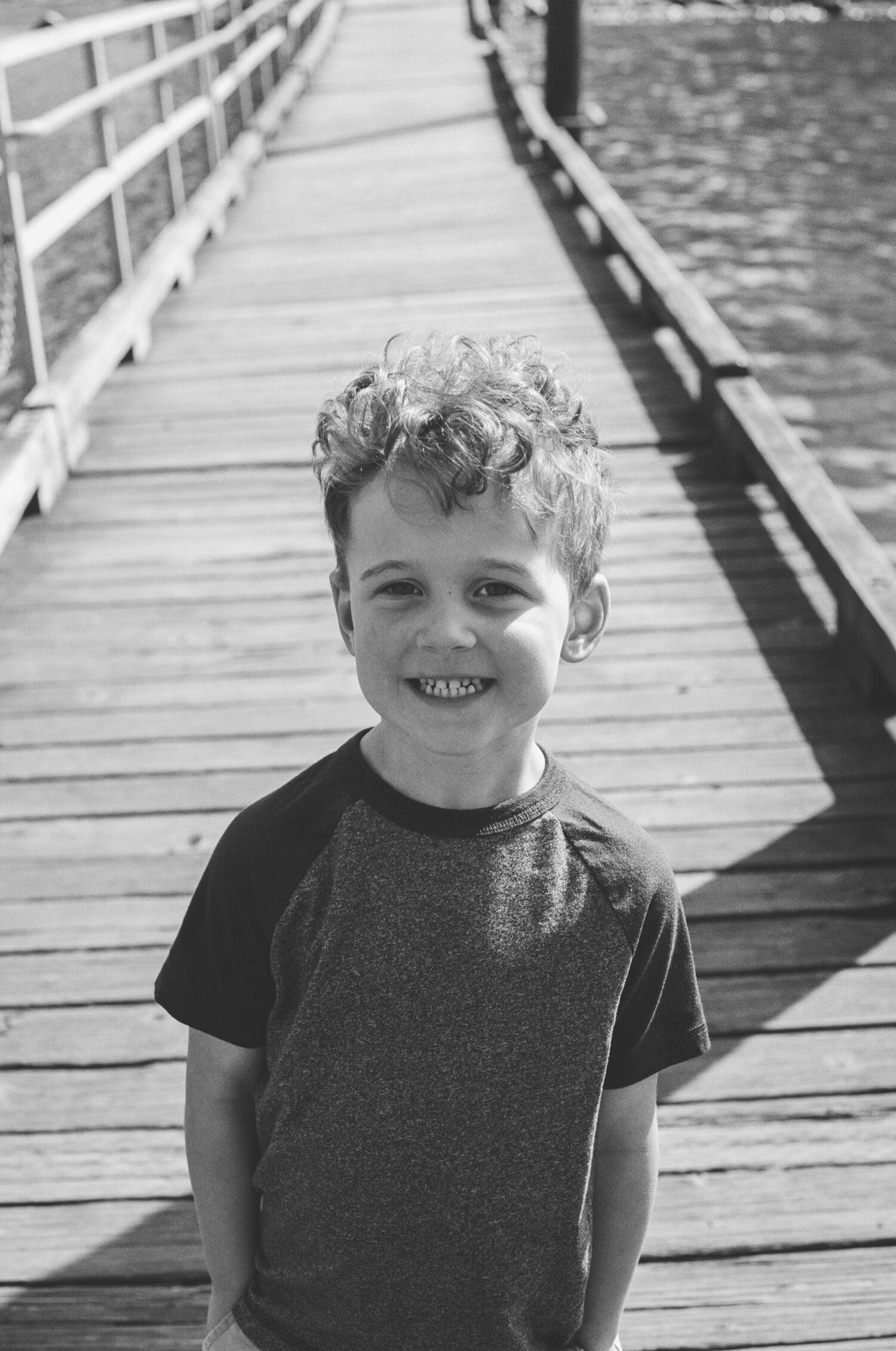 A portrait of a young child standing on a wooden pier with railings over water on a sunny day.