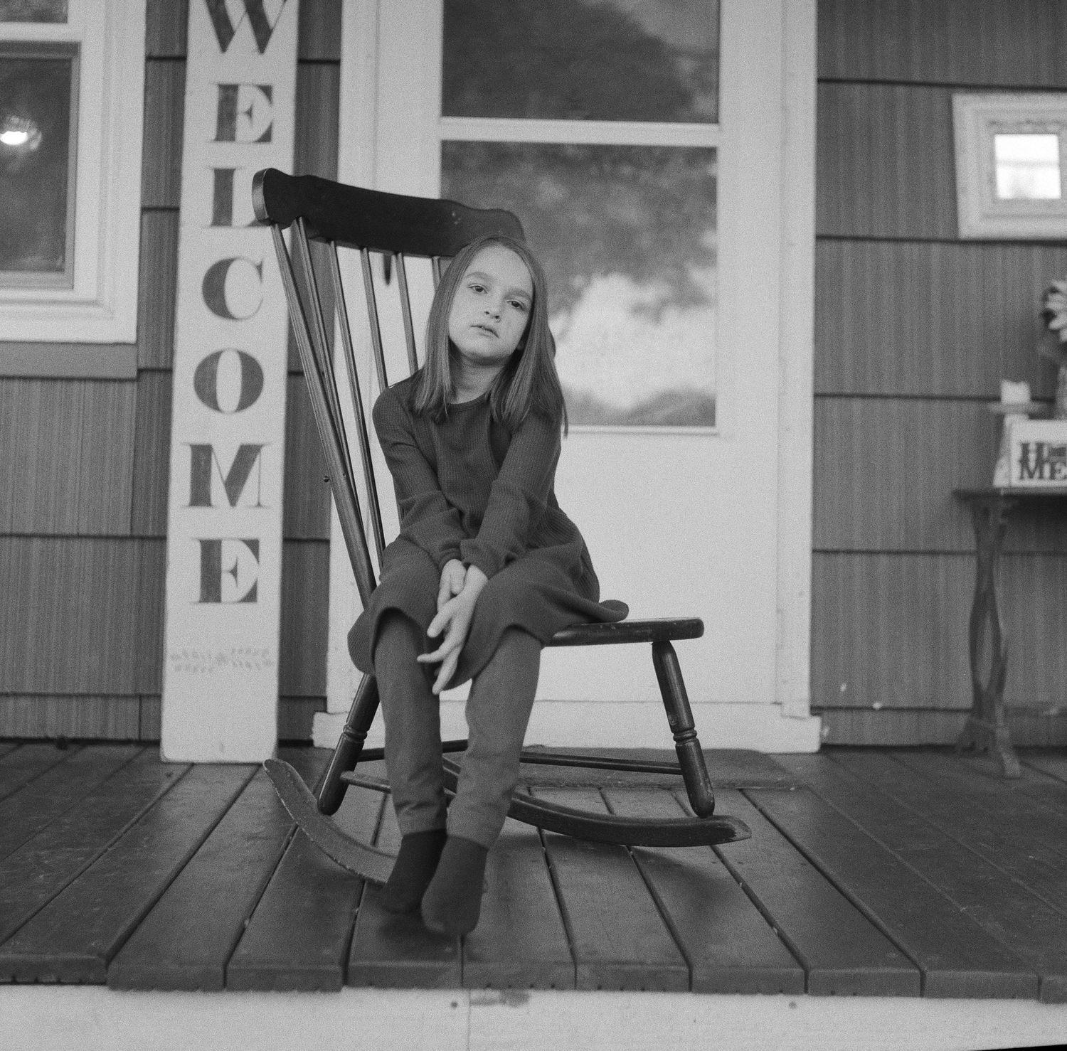 A figure sits on a wooden rocking chair on a porch with a 'Welcome' sign.