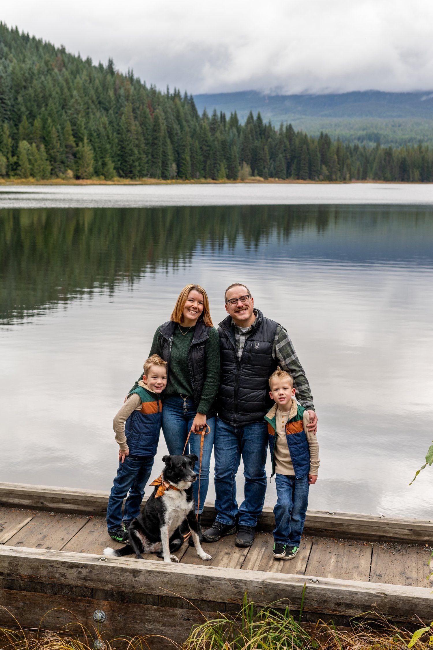 Family with dog poses together on wooden dock by serene mountain lake.