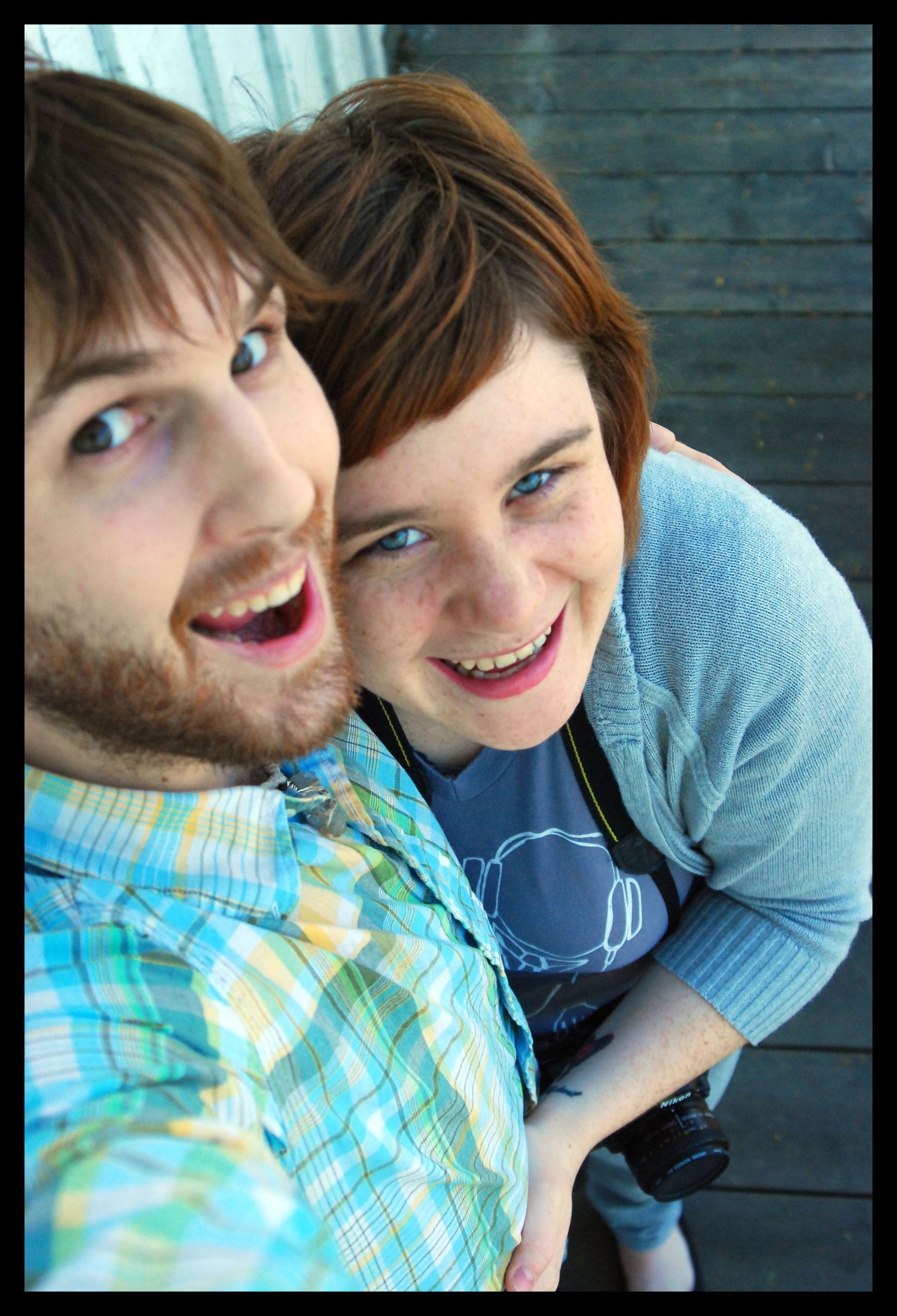 A close-up candid photo of two people smiling and laughing together in front of a teal wooden wall.