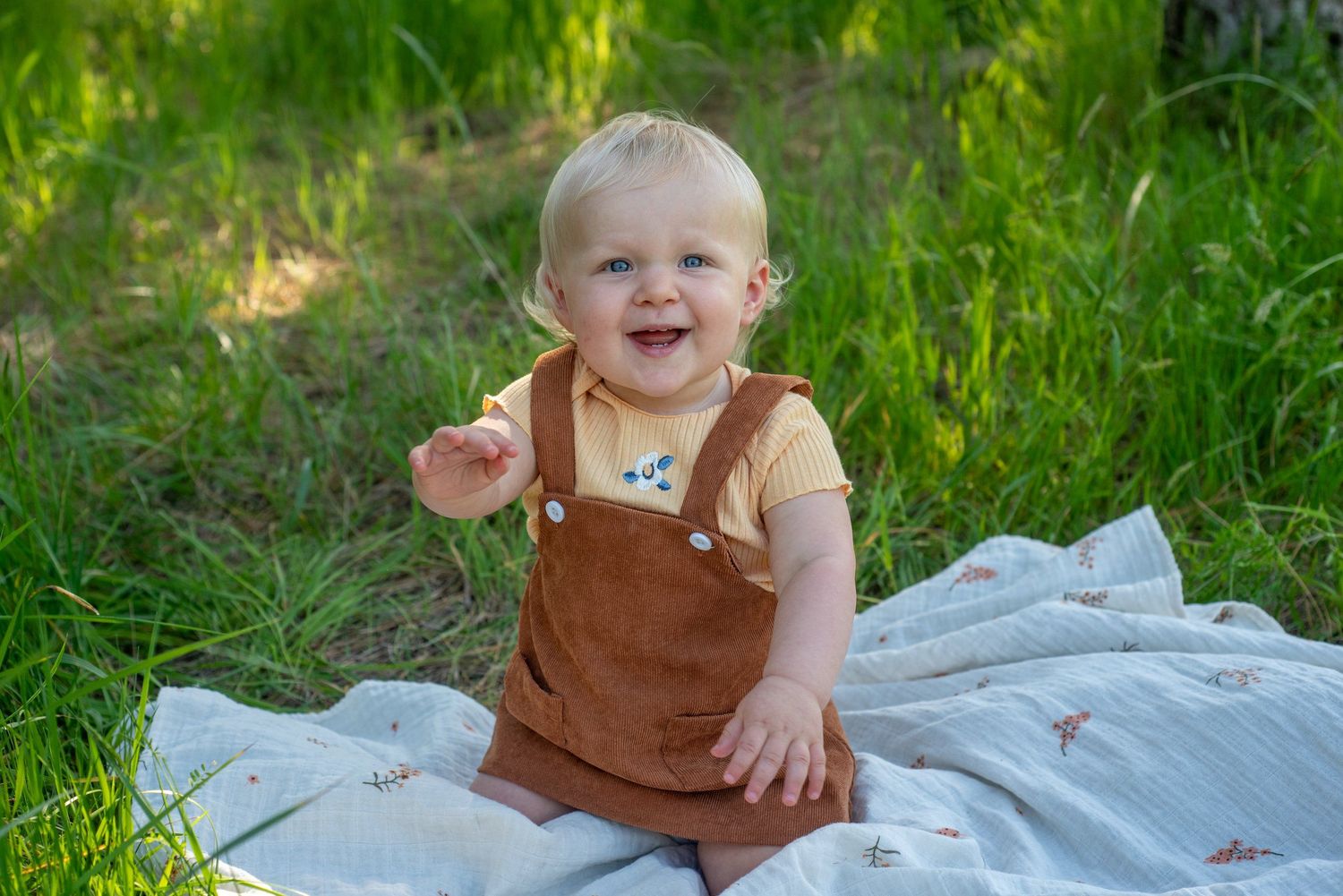 A toddler in brown overalls sits happily on a blanket surrounded by green grass.