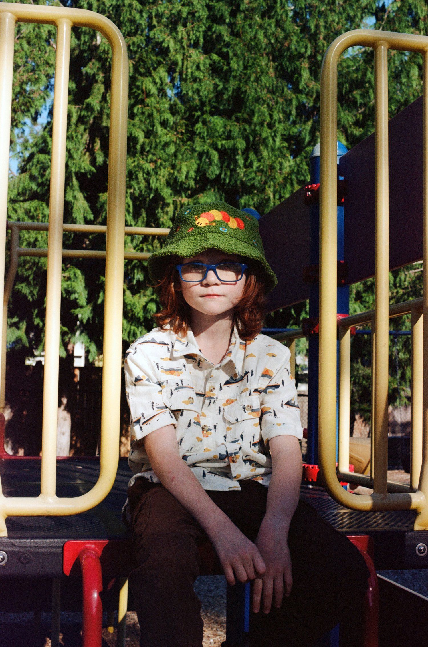 A figure in a patterned shirt and hat sits on playground equipment with greenery in background.