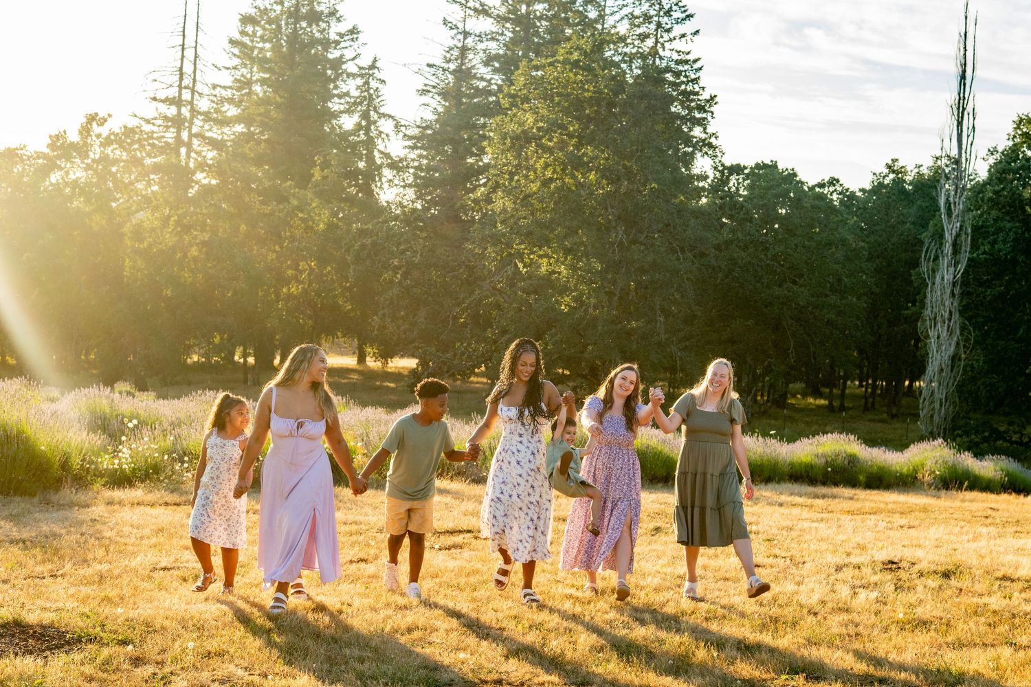 A group of people in summer dresses dance together in a golden sunlit field at sunset with tall trees in the background.