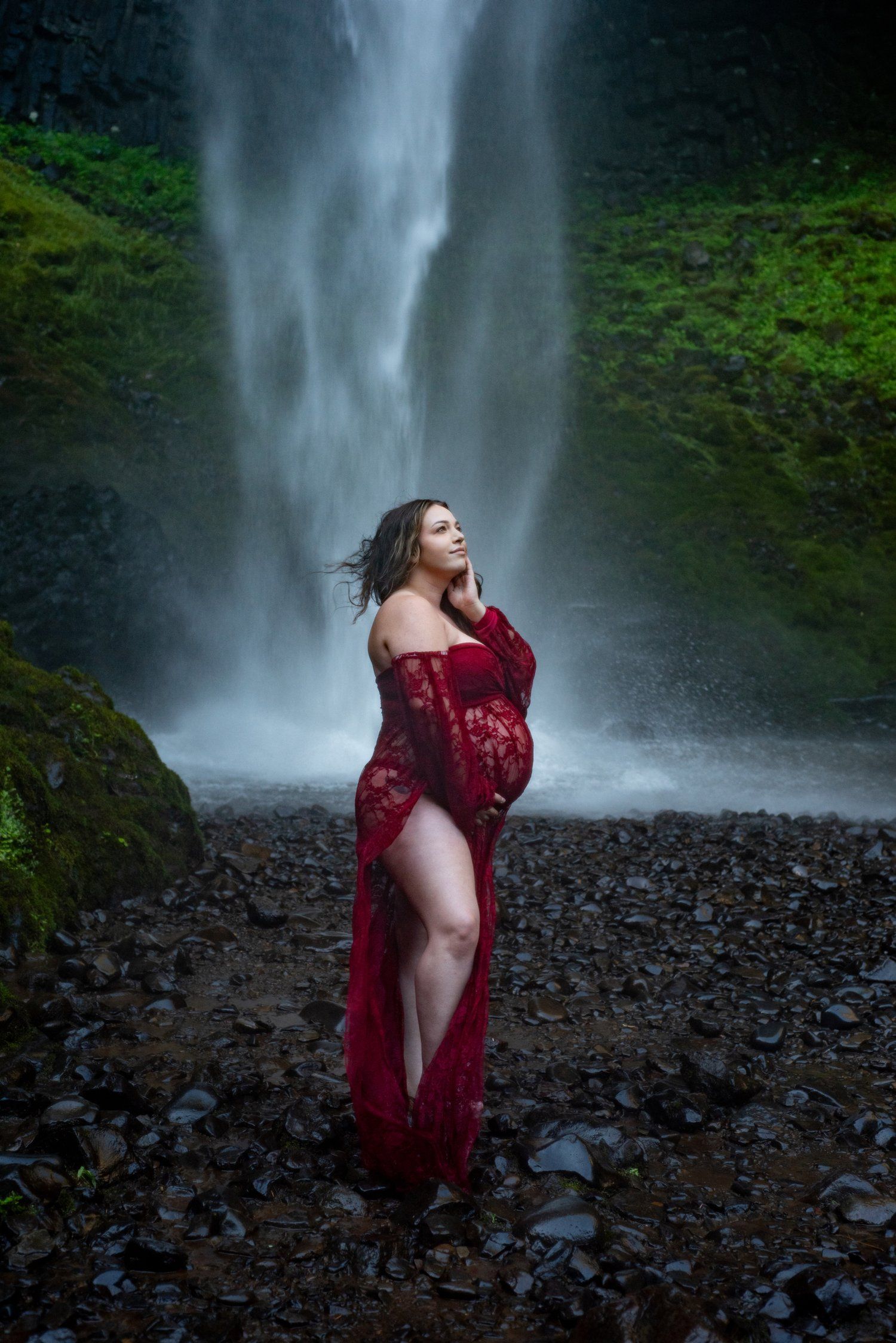 A dramatic maternity photo featuring a red lace dress against a misty waterfall backdrop.