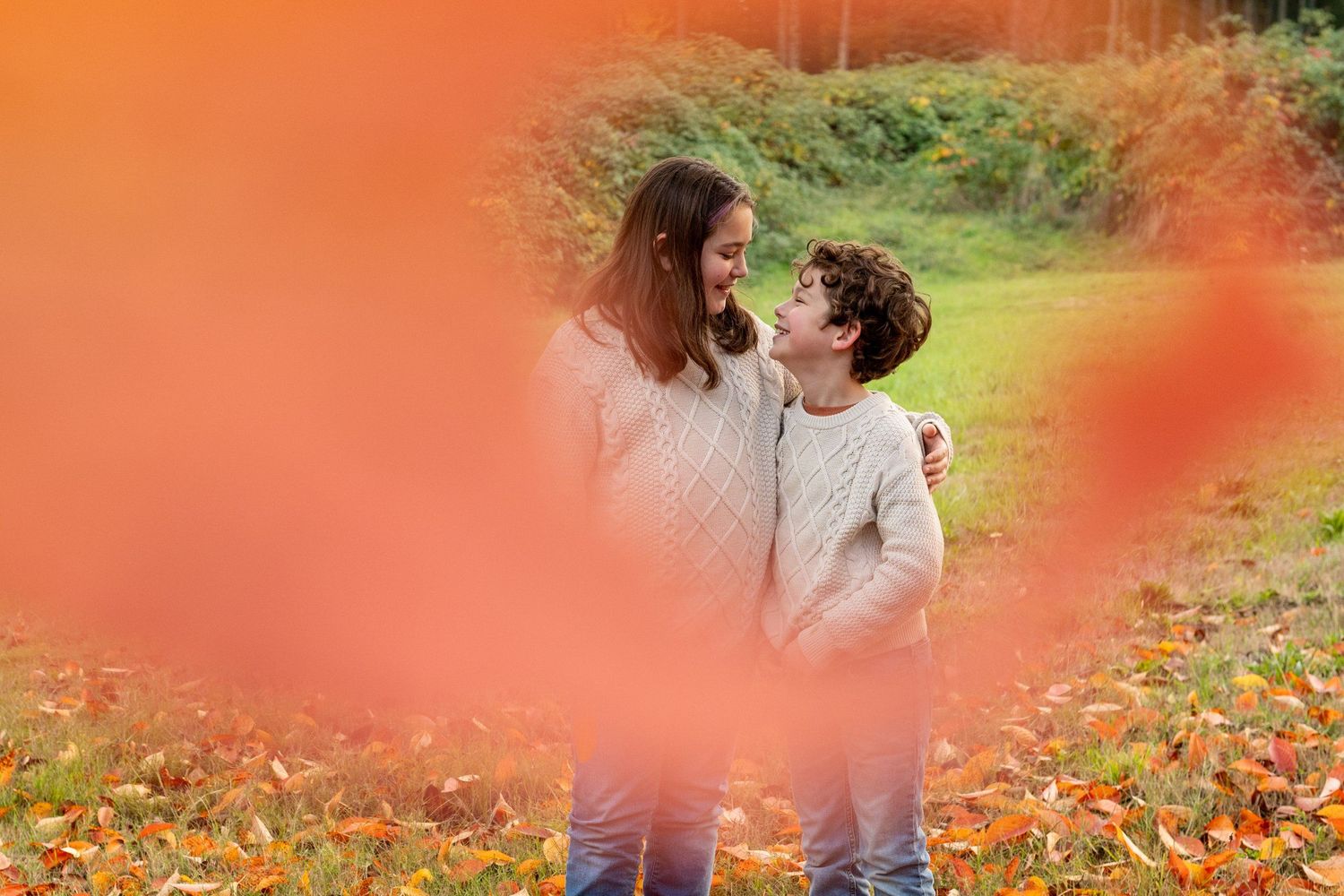 brothers share a moment outdoors among autumn leaves, framed by soft red blurred foreground elements.
