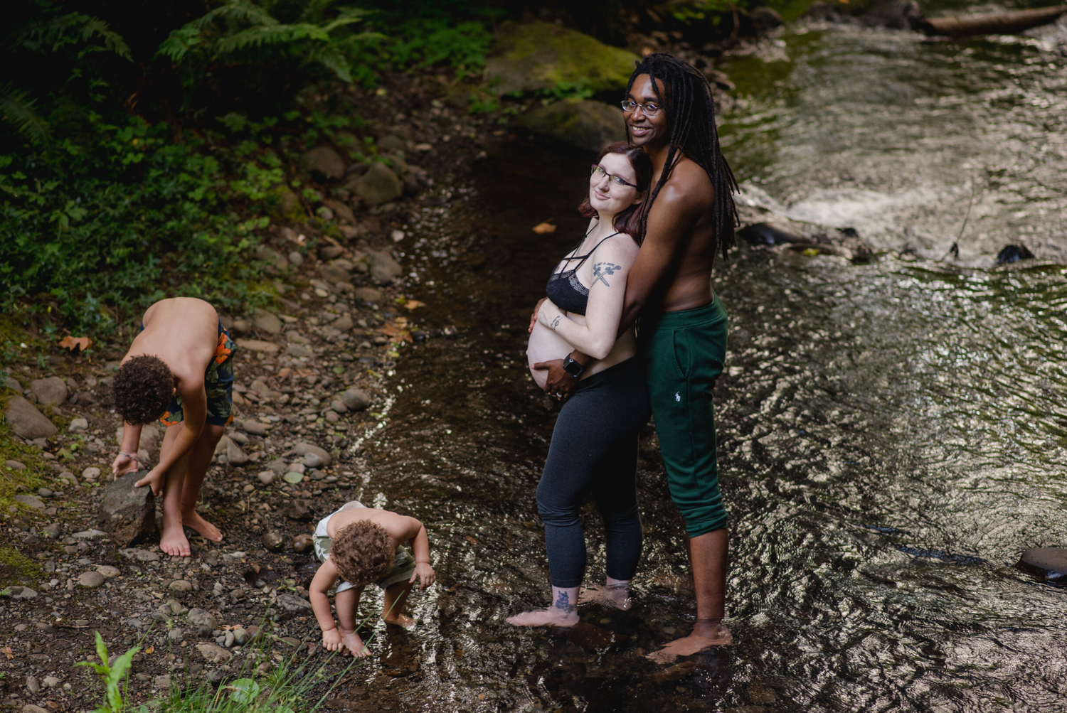People and dogs exploring rocky creek bed in lush forest setting.