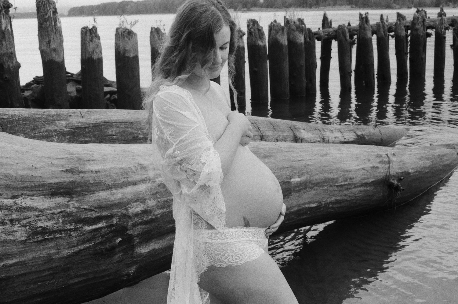 A black and white photo of an expectant mother in a white dress standing by weathered pier pilings.