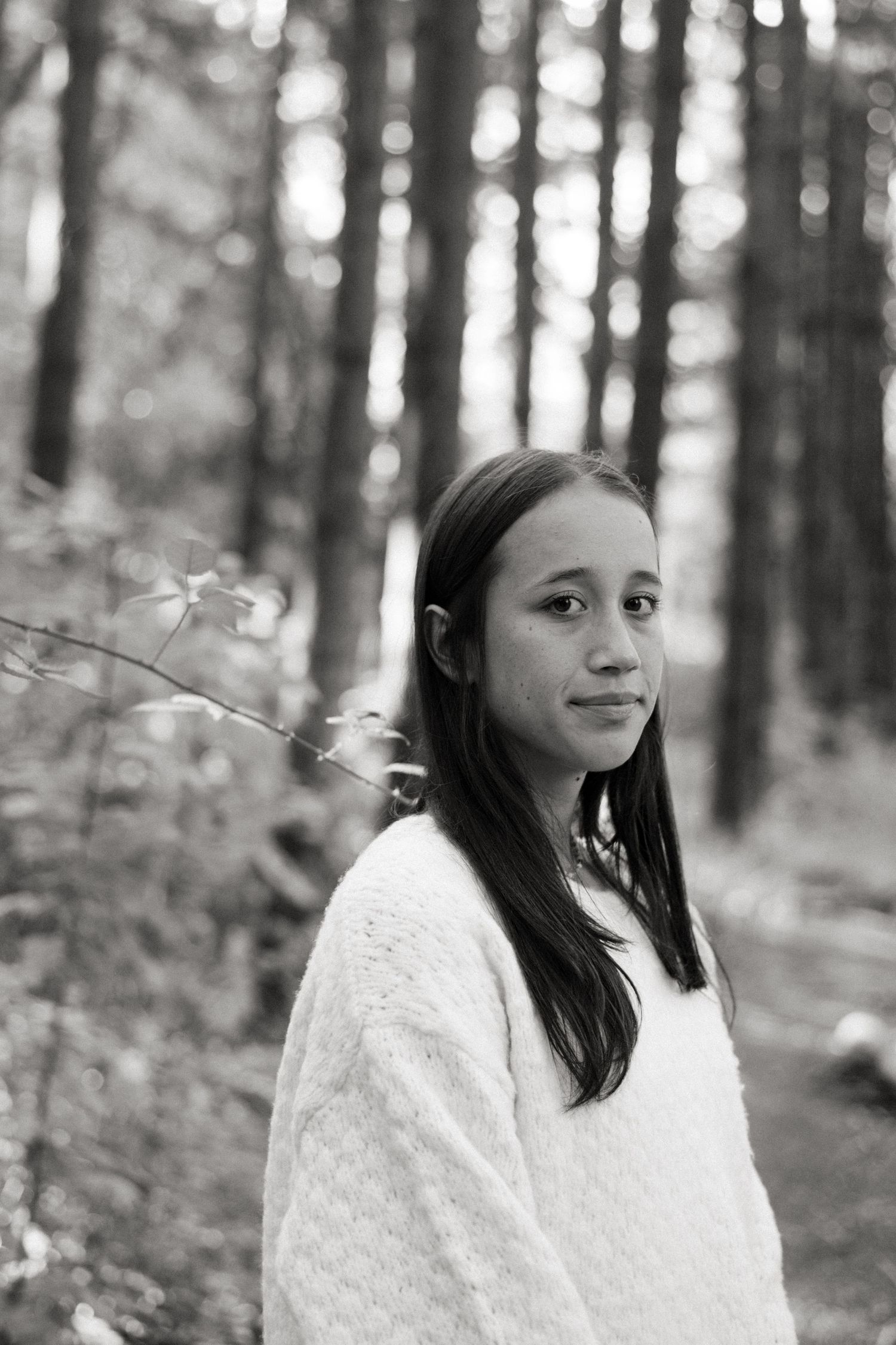 A young person in a cozy white sweater stands in a peaceful forest setting captured in black and white photography.