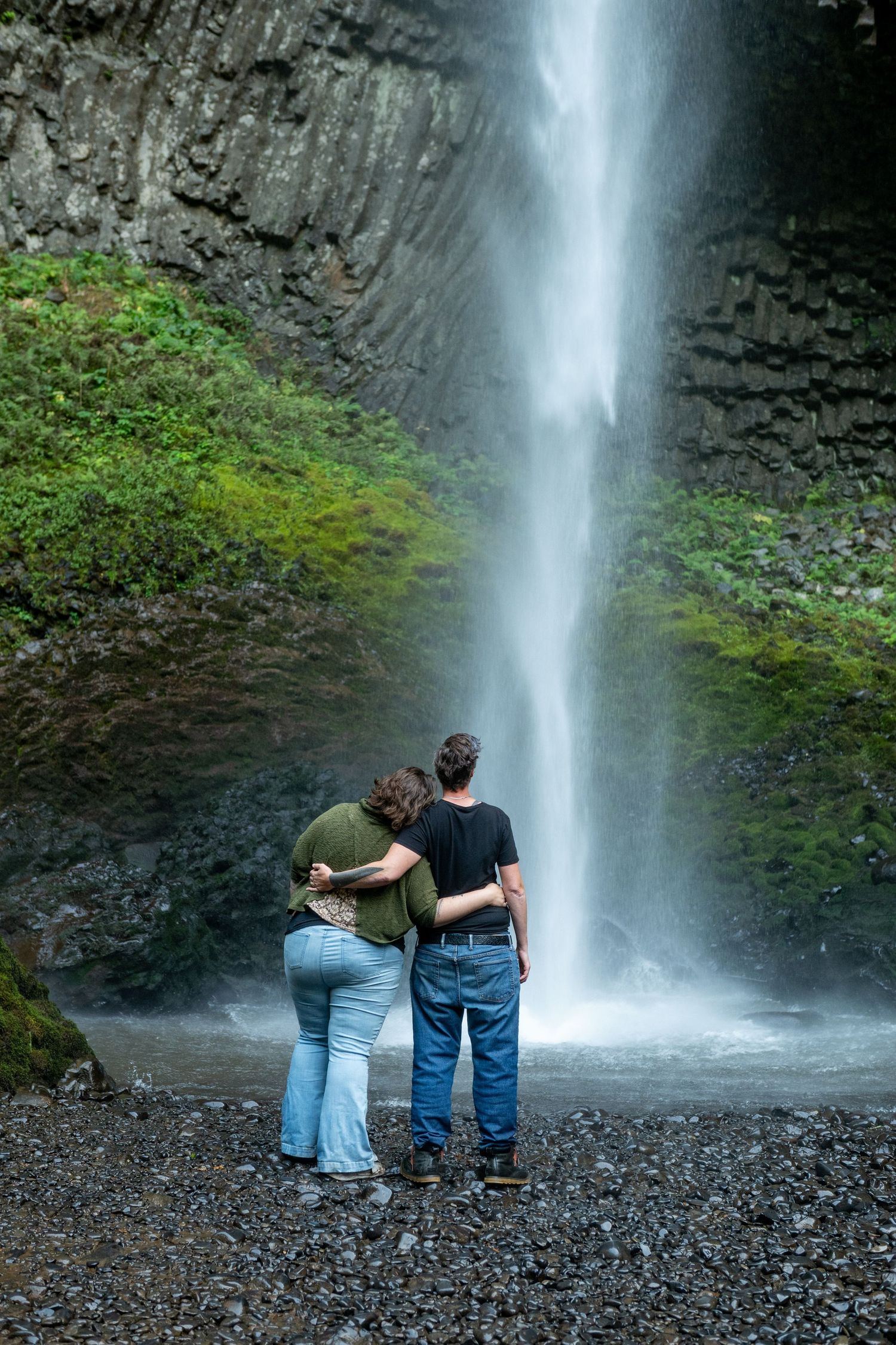 Two people stand close together while admiring a tall cascading waterfall among rocky cliffs and green moss.