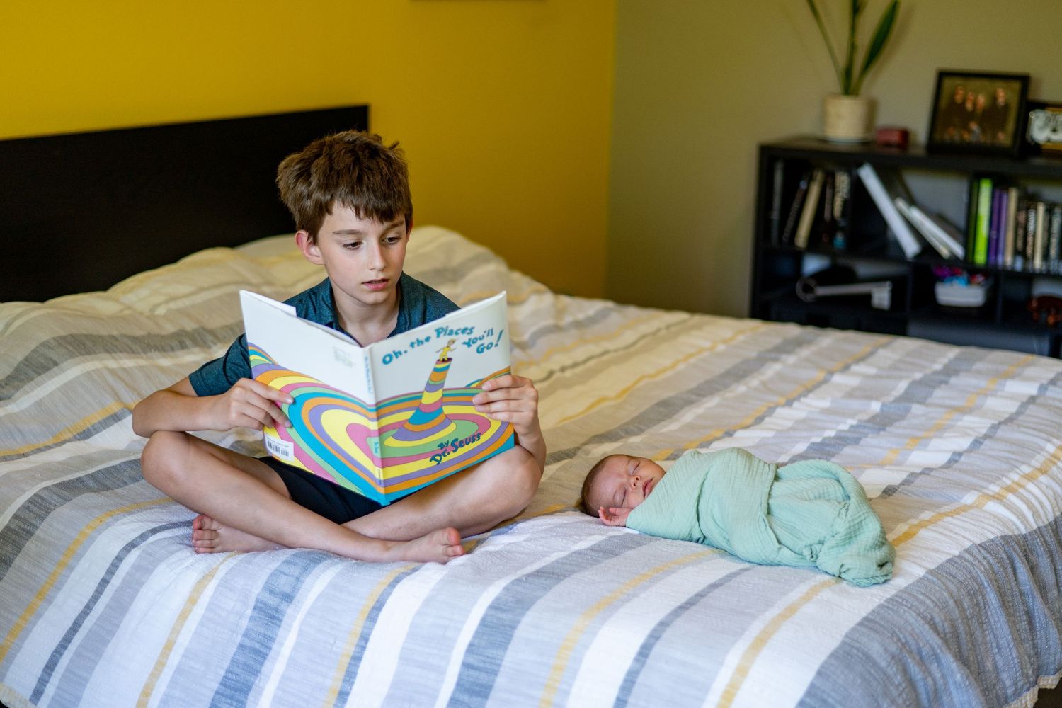 Reading a book to baby on a striped bedspread in a yellow-walled bedroom.