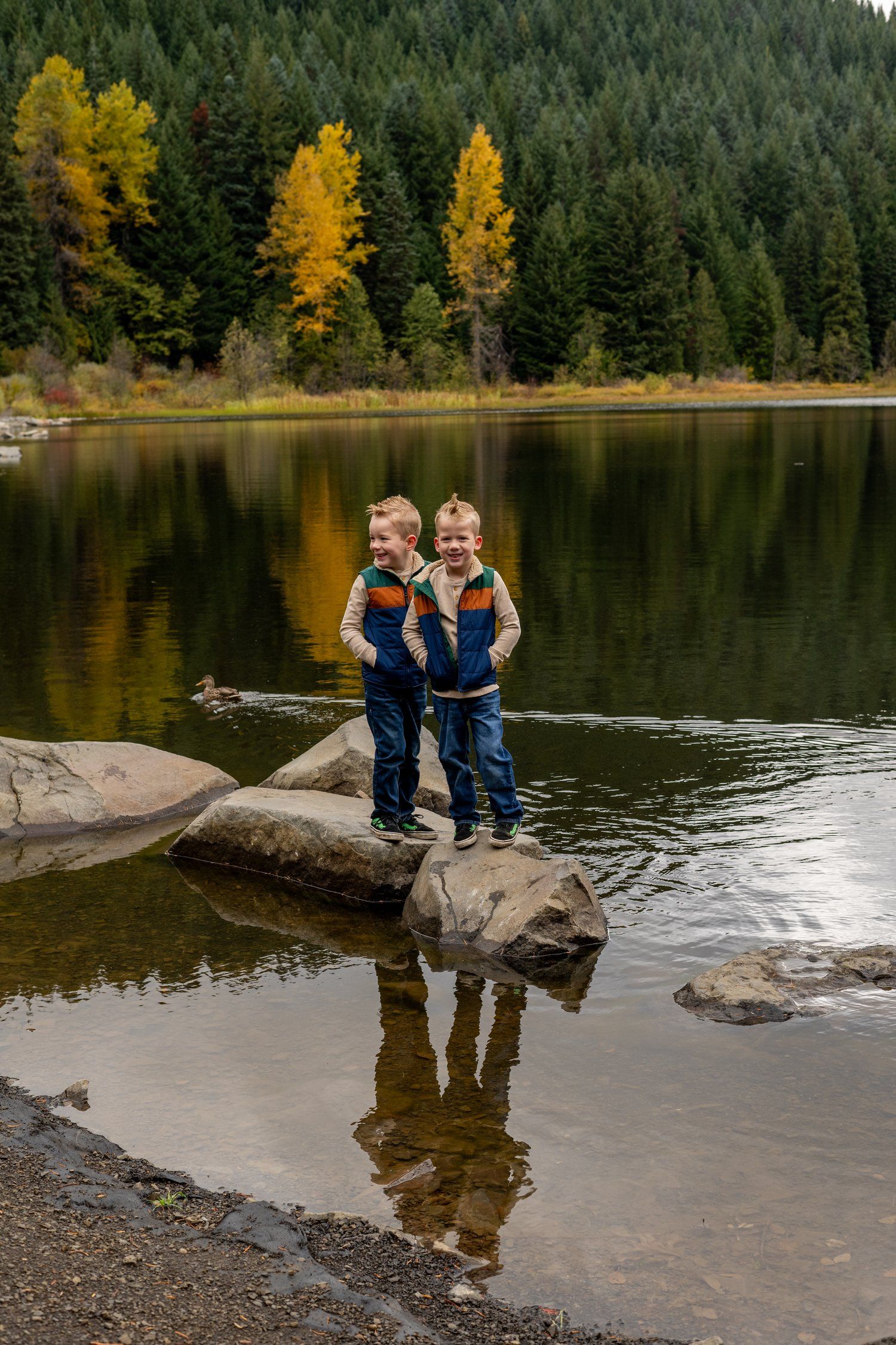 Two children stand on rocky shore of mountain lake surrounded by autumn trees.