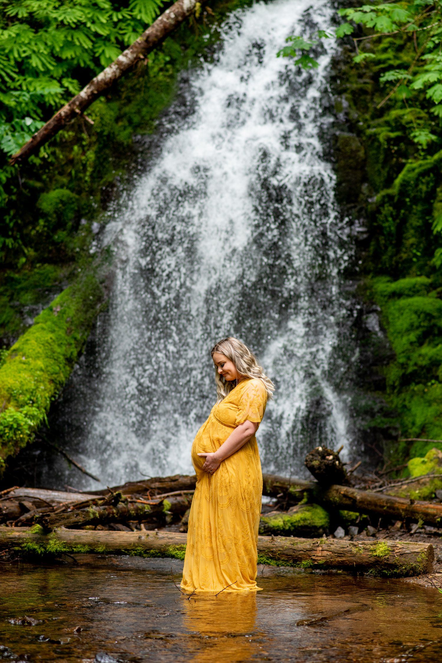 Expecting mother in flowing yellow dress stands in front of majestic waterfall surrounded by lush greenery.