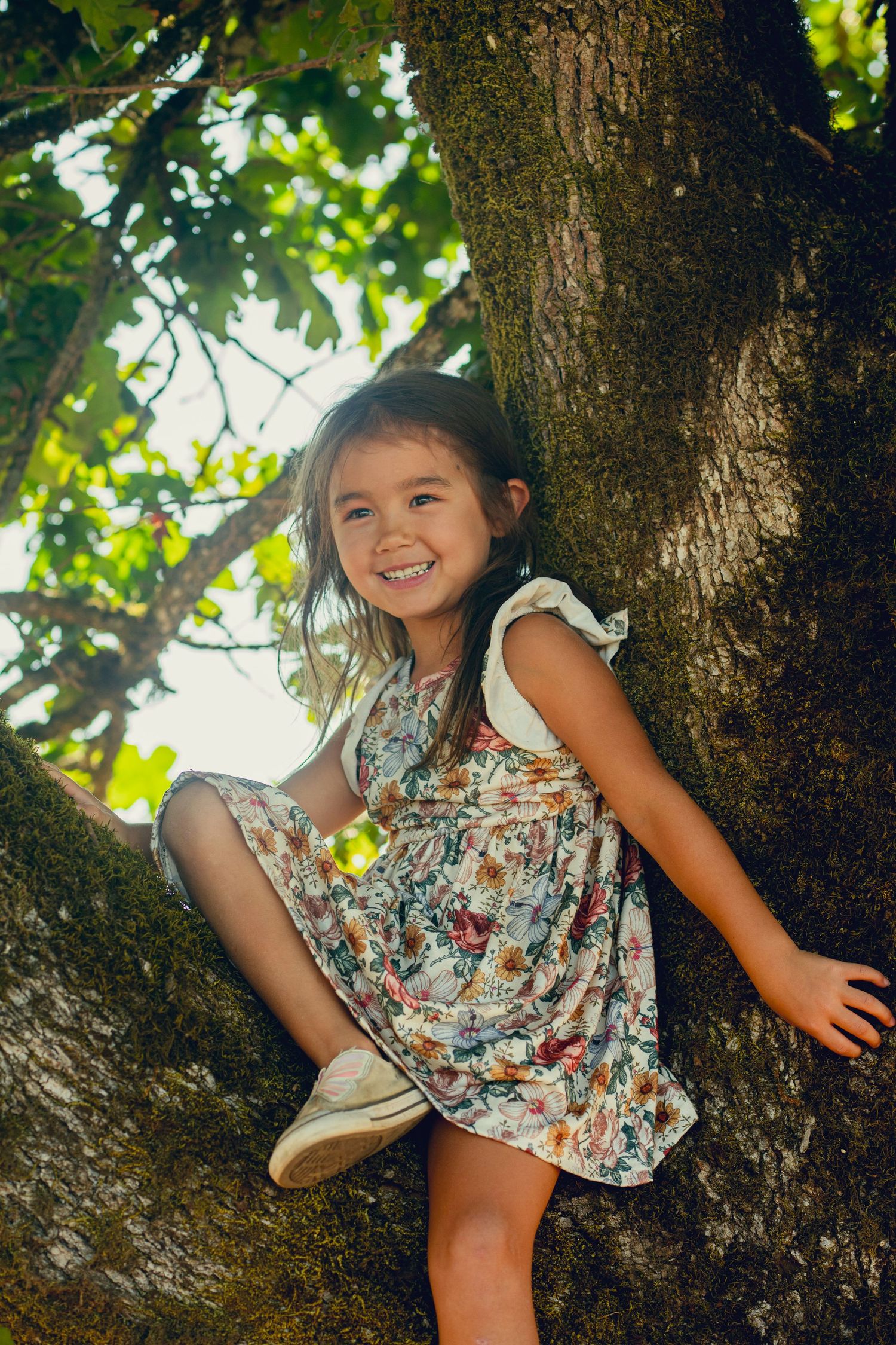 Child in floral dress plays joyfully on a tree branch on a sunny summer day.